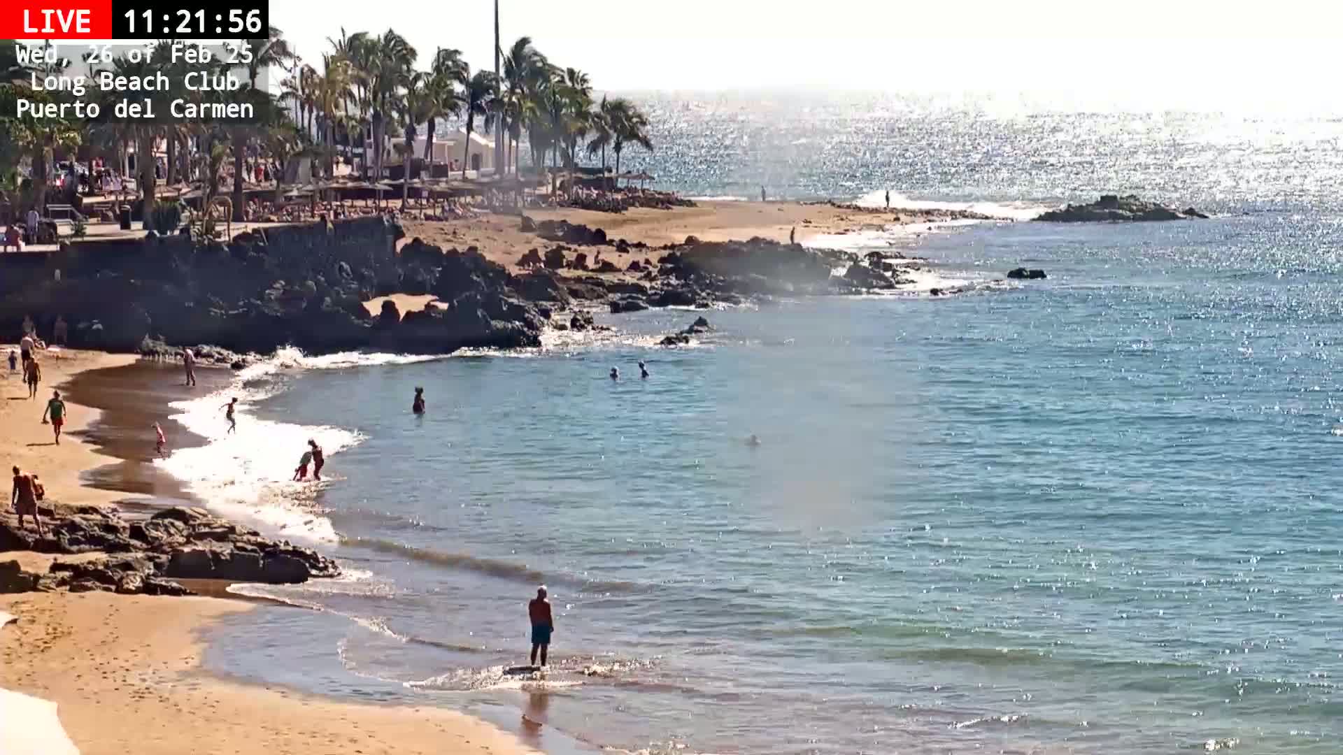 A sandy beach with calm, clear water and several people swimming and sunbathing under a sunny sky.