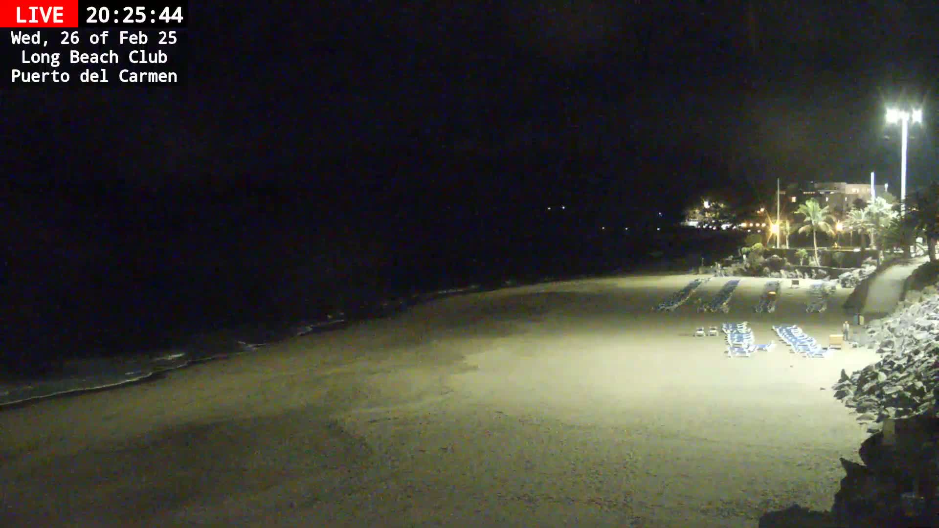 A mostly empty sandy beach at night, illuminated by artificial lights, with rows of lounge chairs and a calm ocean visible in the background.