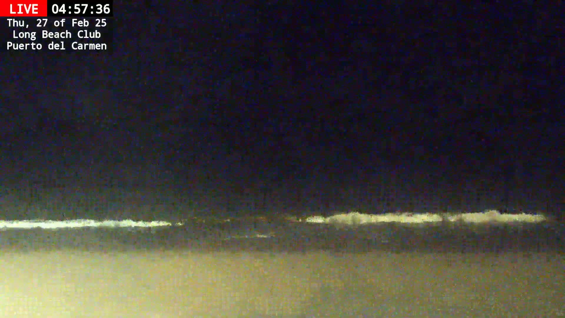 A dark, nighttime beach scene shows a faint line of waves breaking on the shore under a dark sky.