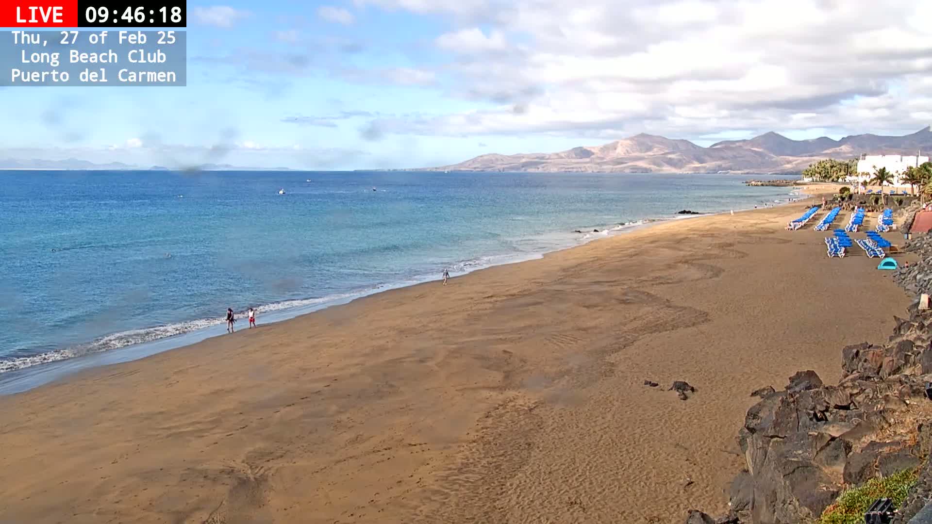 A sandy beach with calm ocean waves stretches towards distant mountains under a partly cloudy sky.