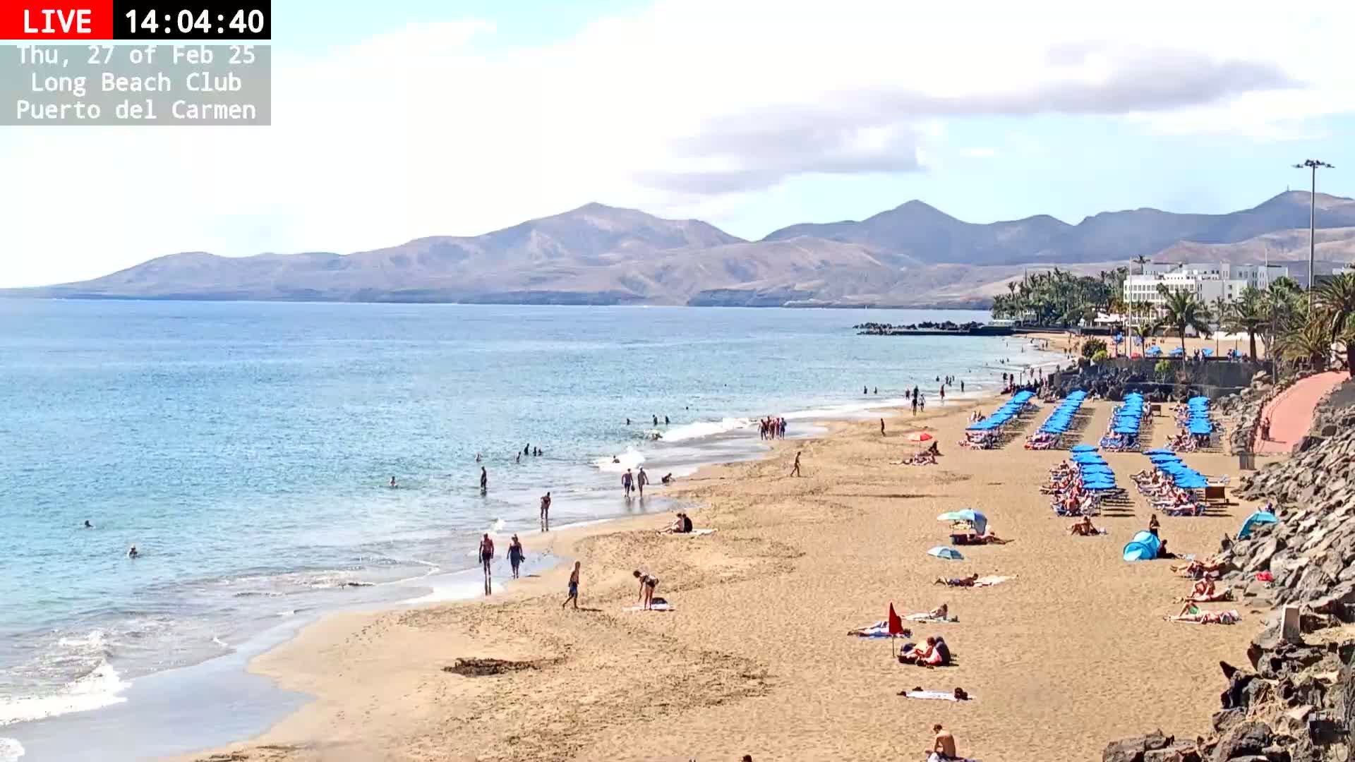 A sandy beach with numerous blue umbrellas and sunbathers stretches towards a calm ocean under a partly cloudy sky, with mountains visible in the background.