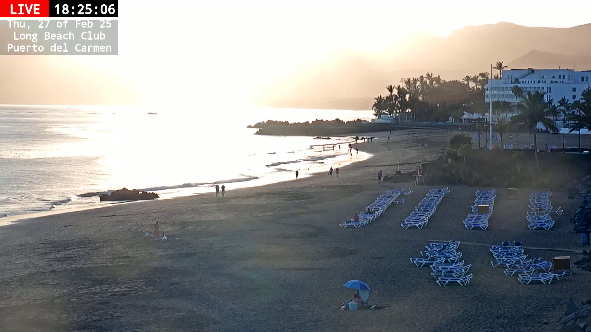 A sandy beach with scattered people, rows of empty lounge chairs, and a calm ocean under a hazy sunset.
