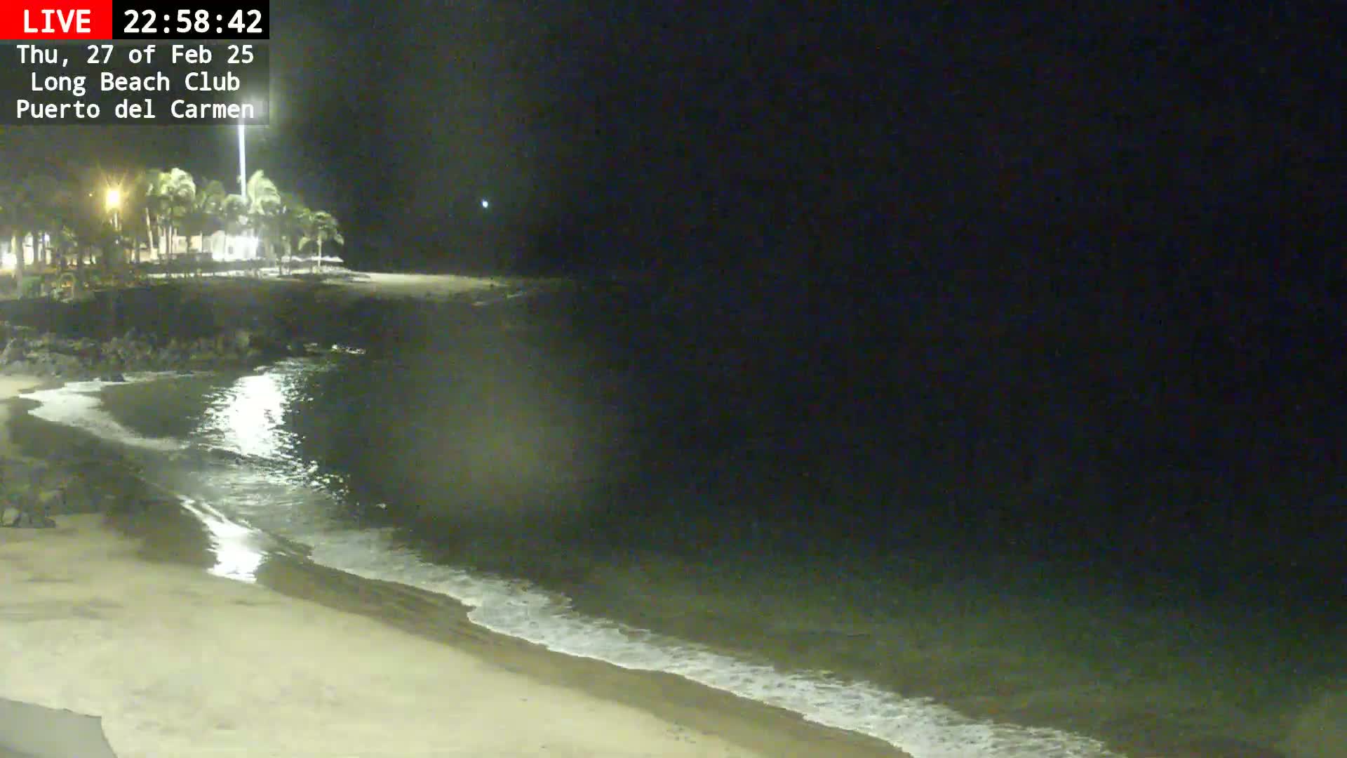 A dark, nighttime beach scene shows a sandy shore with gentle waves lapping at the edge of the water, under a clear, dark sky.