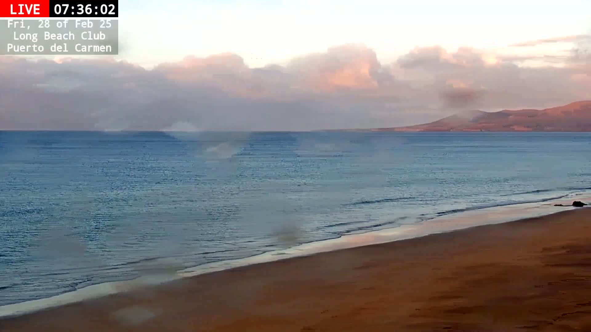 A sandy beach meets a calm ocean under a partly cloudy, pinkish sky with a distant mountain range visible on the horizon.