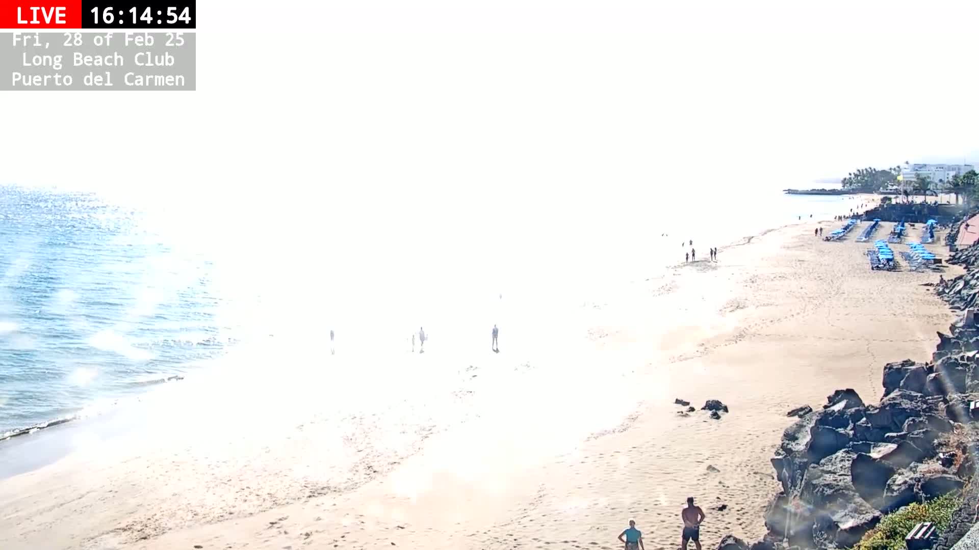 A mostly sunny beach scene shows a sandy shore with a few people and rows of beach chairs, bordering a calm ocean under a bright sky.