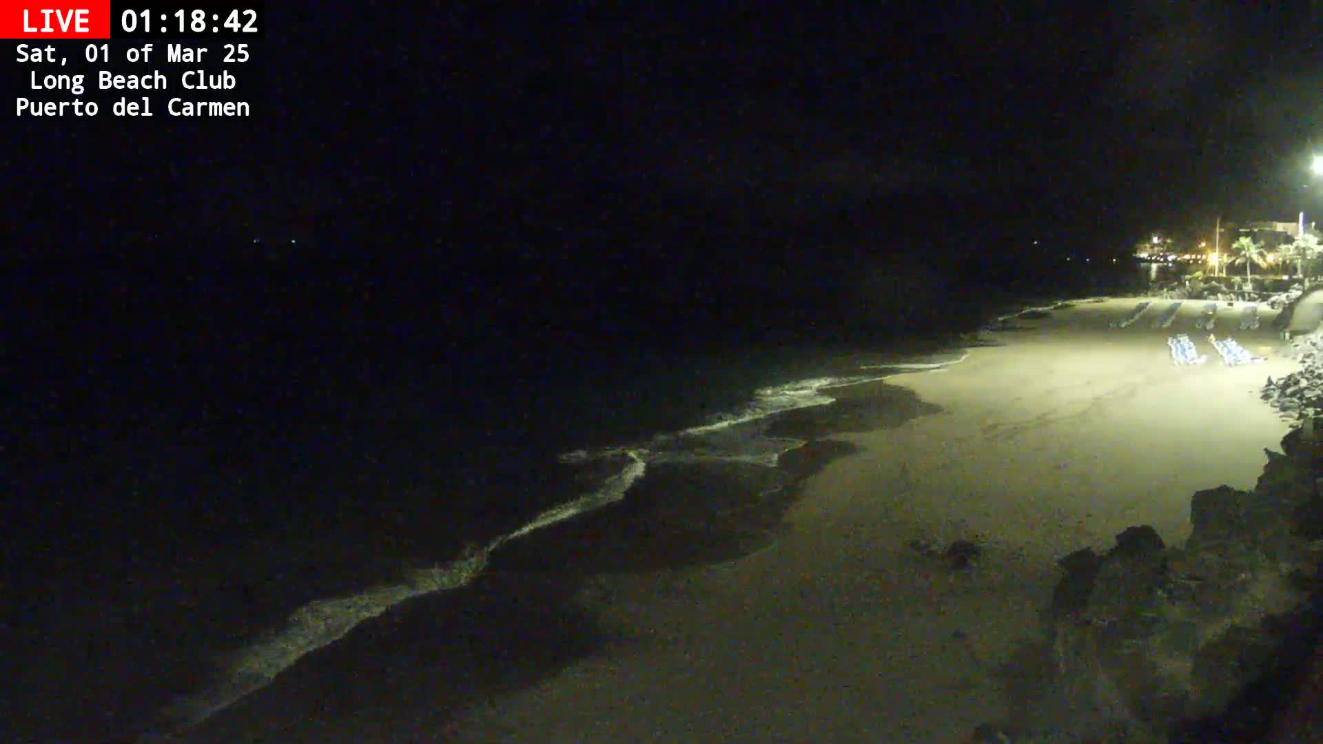 A dark, nighttime beach scene shows gently lapping waves on a sandy shore, with some beach chairs visible in the distance under artificial light.
