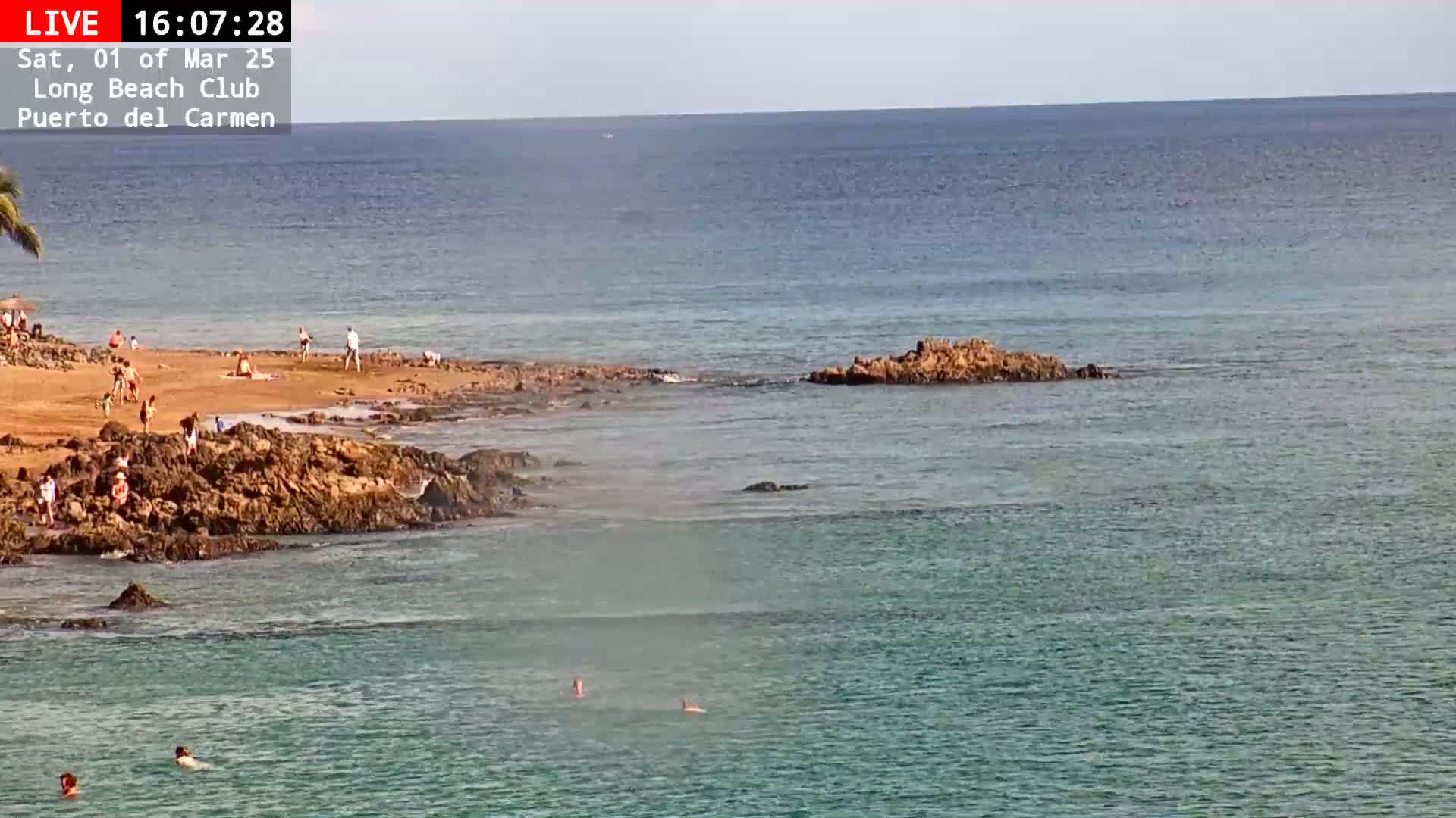 A sandy beach with people sunbathing and swimming in calm, clear turquoise water near rocky outcrops under a mostly sunny sky.