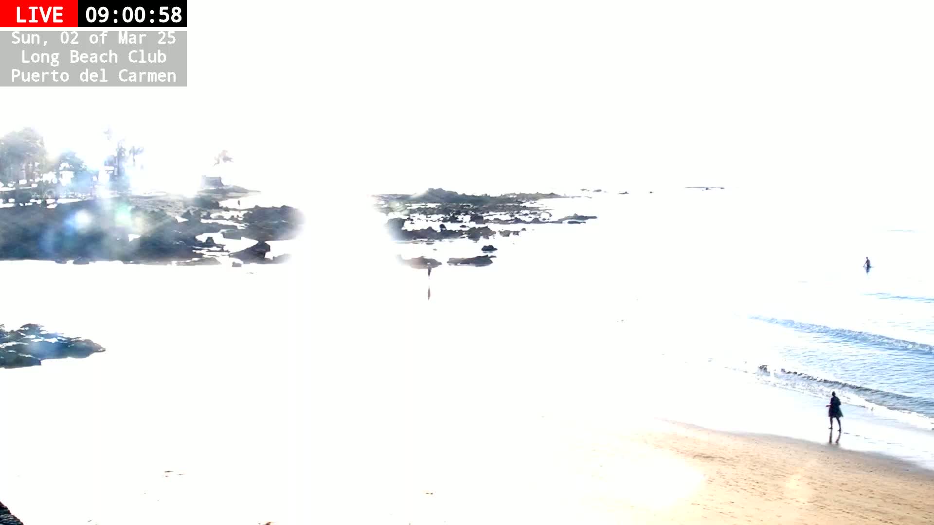 A mostly sunny beach scene shows a sandy shore with a few people walking, dark rocks in the shallow water, and calm waves rolling onto the beach.