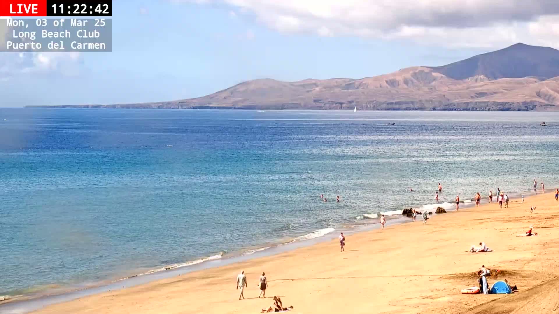 A sandy beach with scattered people swimming and relaxing, under a partly cloudy sky, stretches to a calm ocean and distant mountains.
