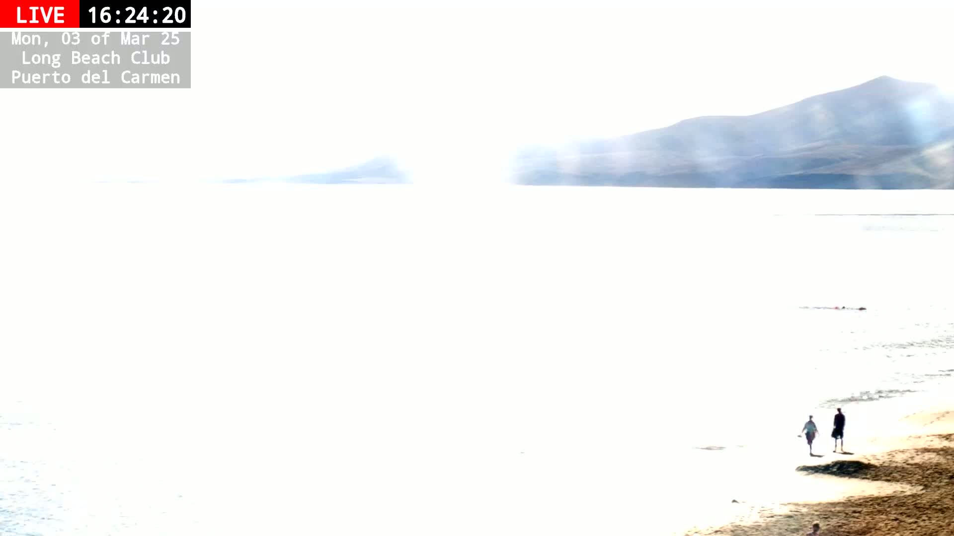 Two people walk along a sandy beach toward the viewer, with a hazy, bright sunlit ocean and distant mountains in the background.