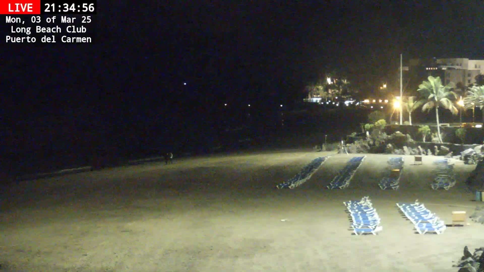 A dark beach at night with rows of empty lounge chairs, a few people in the distance, and palm trees illuminated by lights in the background.