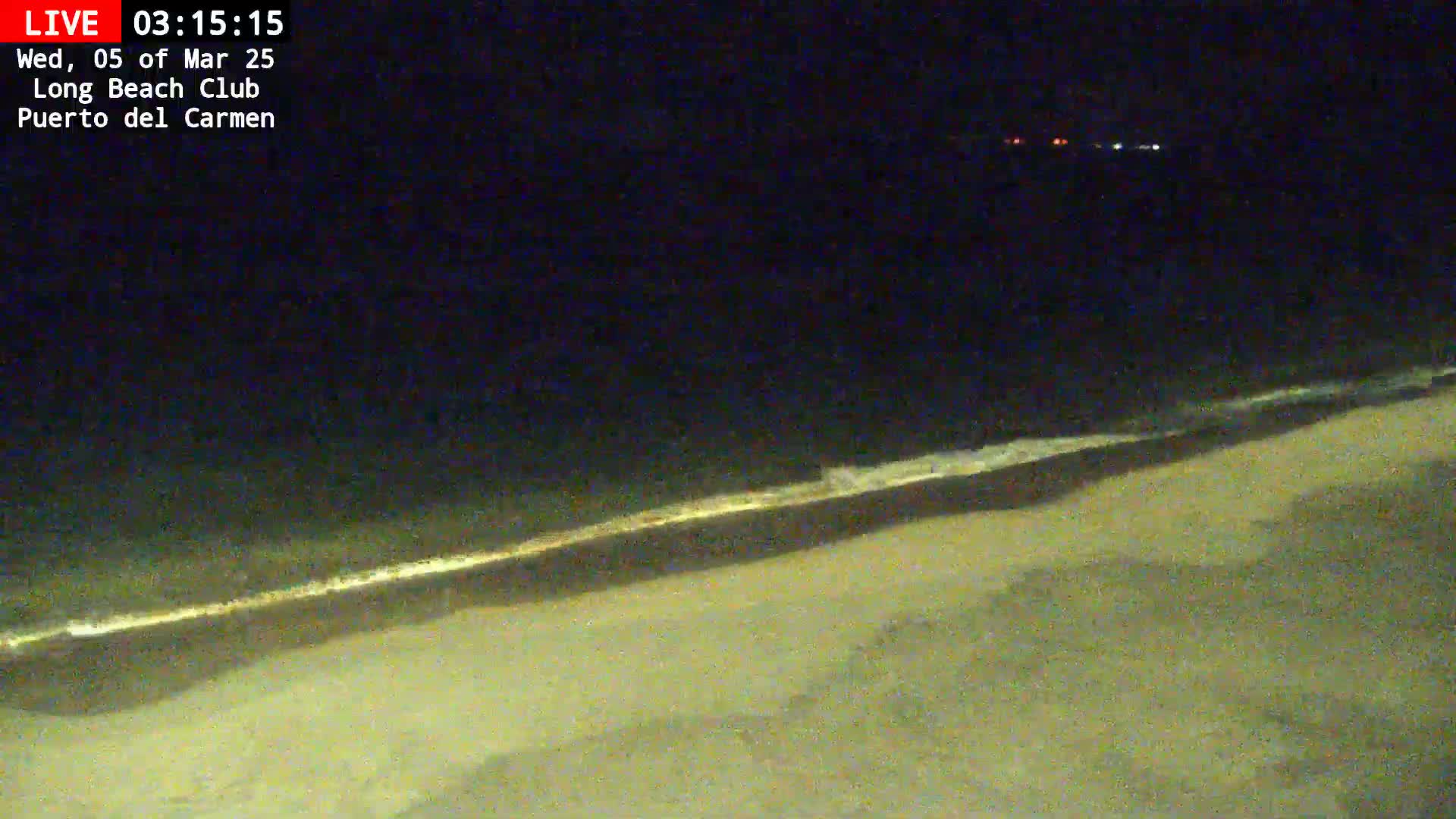 A dark beach at night shows a line of small waves lapping the shore under a clear, dark sky.