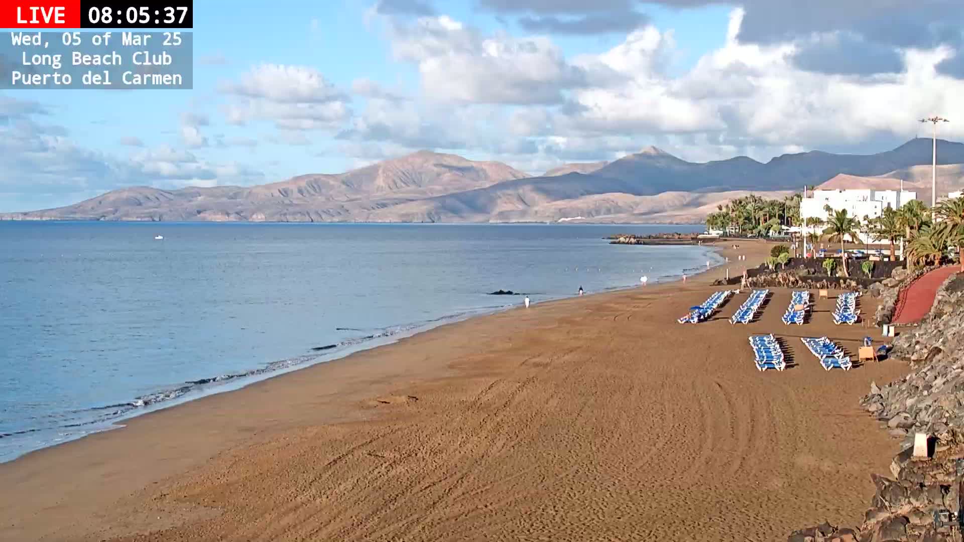 A sandy beach with rows of empty lounge chairs stretches toward a calm ocean under a partly cloudy sky, backed by distant mountains.