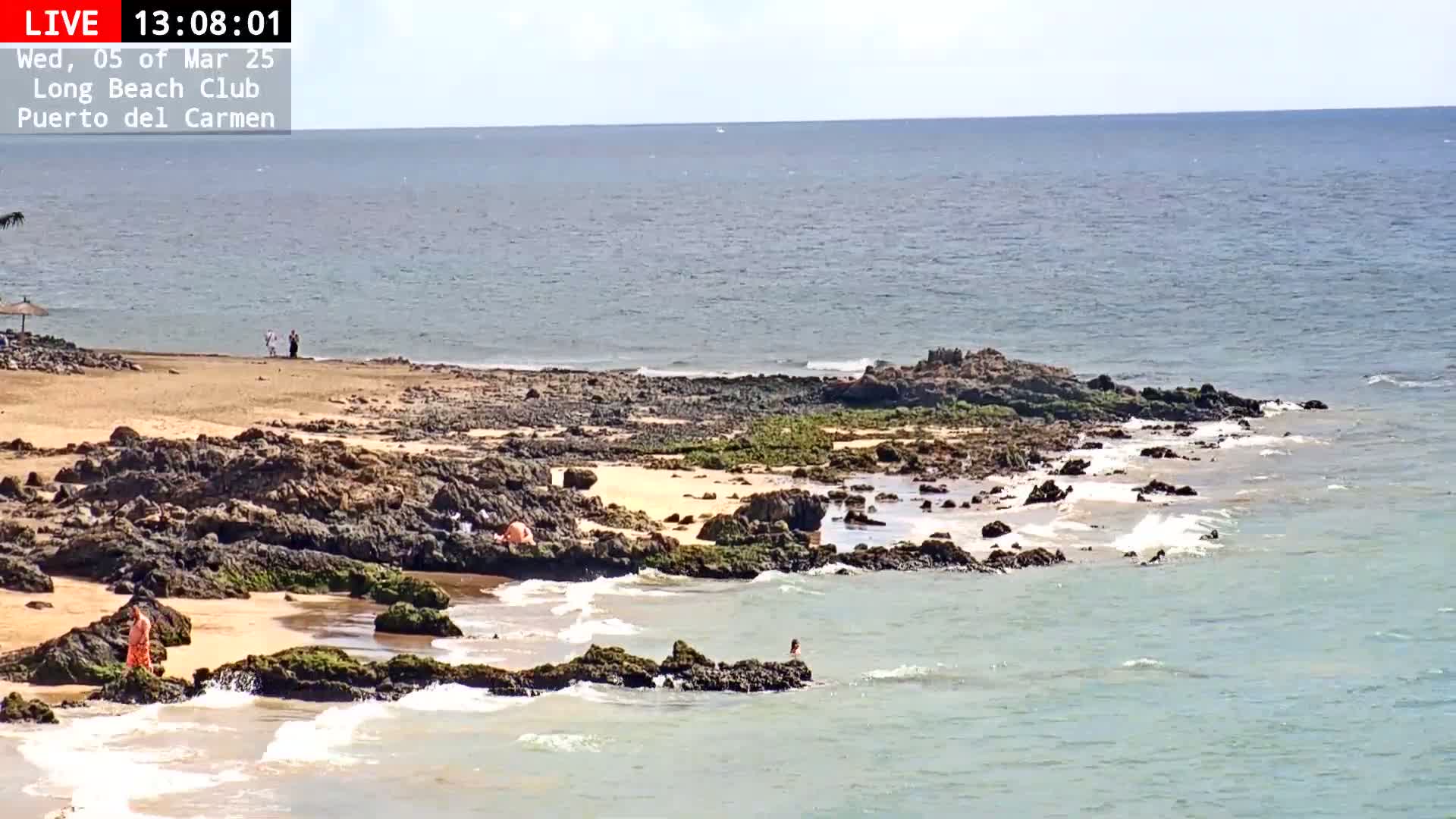 A rocky shoreline with a sandy beach, calm ocean waves, and a few people are visible under a mostly sunny sky.