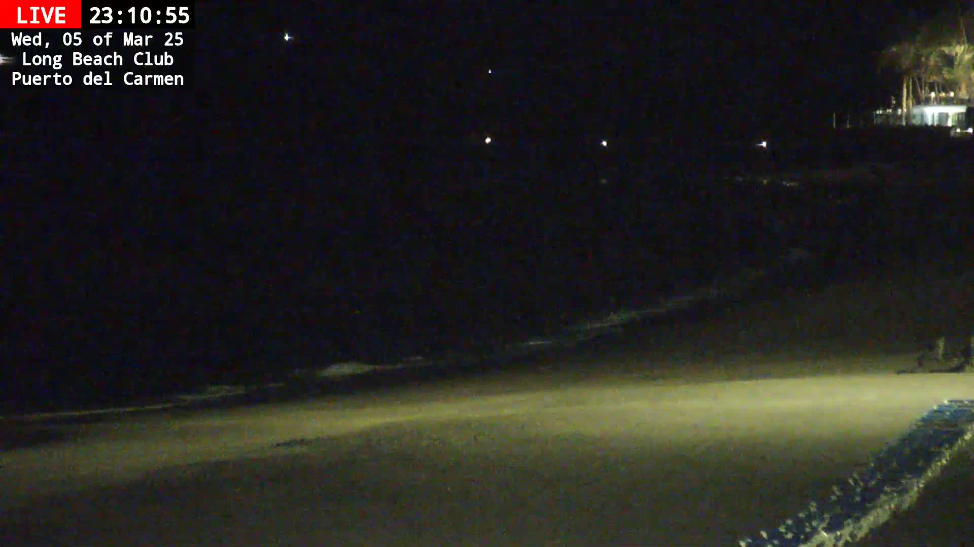 A dark, nighttime beach scene shows gently lapping waves on a sandy shore, with distant lights visible in the darkness.