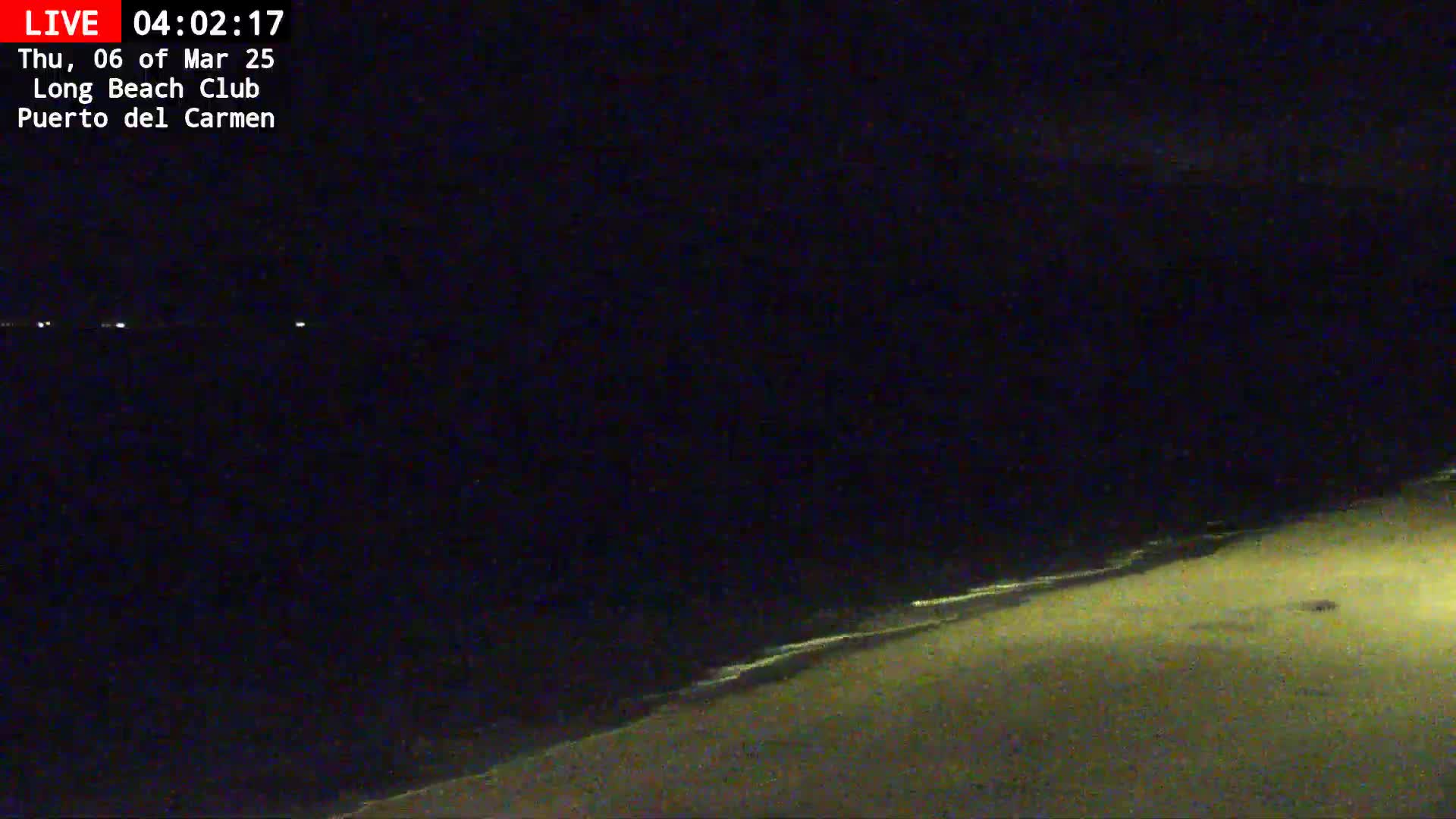 A dark, nighttime beach scene shows a sandy shore with gently lapping waves under a mostly clear sky.