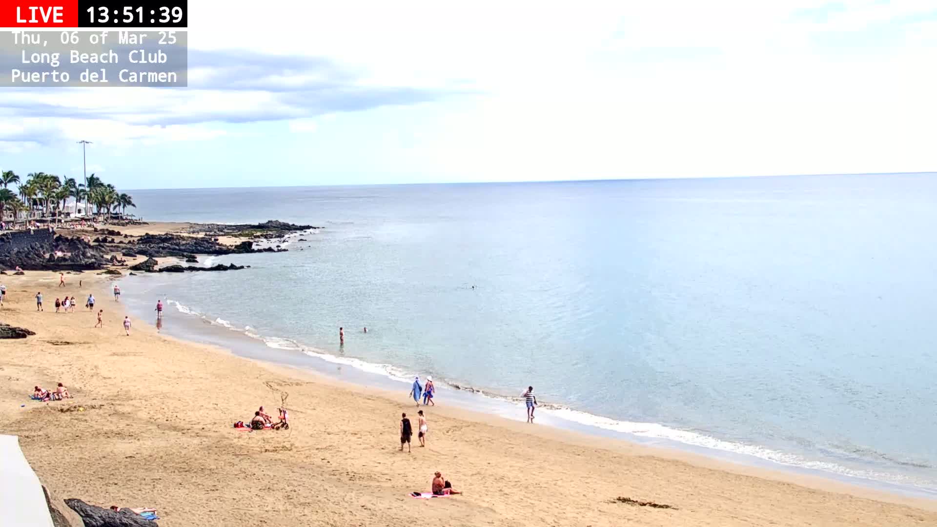 A sandy beach with a calm, light-blue ocean under a partly cloudy sky has several people enjoying the sun and water.
