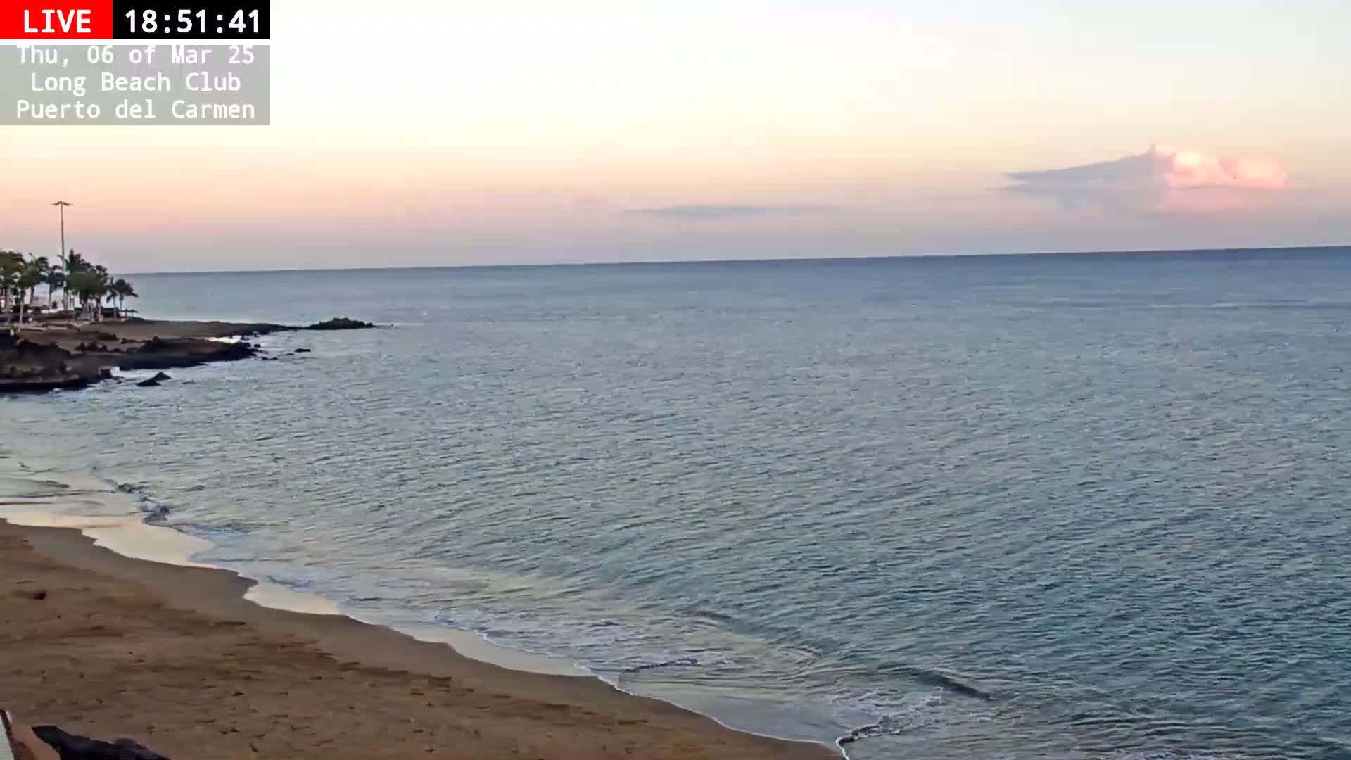 A sandy beach meets a calm ocean under a pale pink and orange sunset sky.