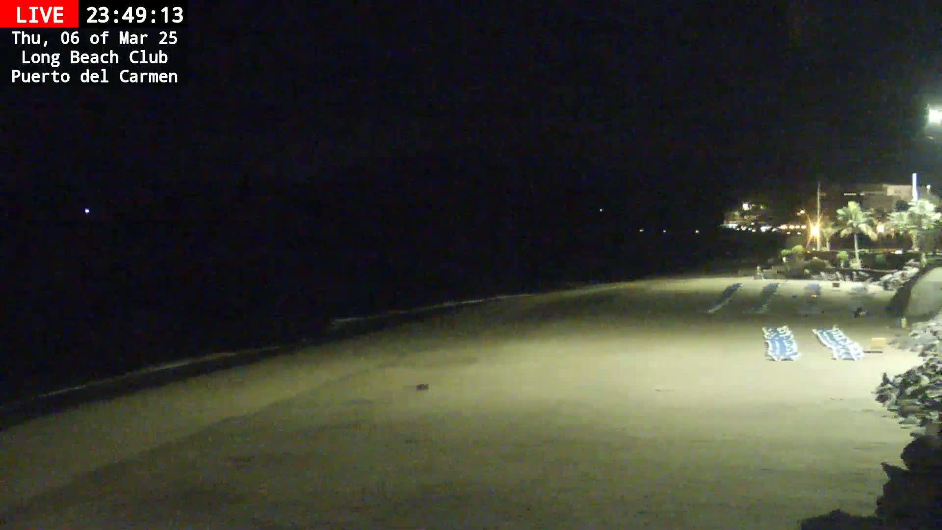 A dark, sandy beach at night is sparsely populated with folded beach chairs, and faintly lit buildings are visible in the distance.