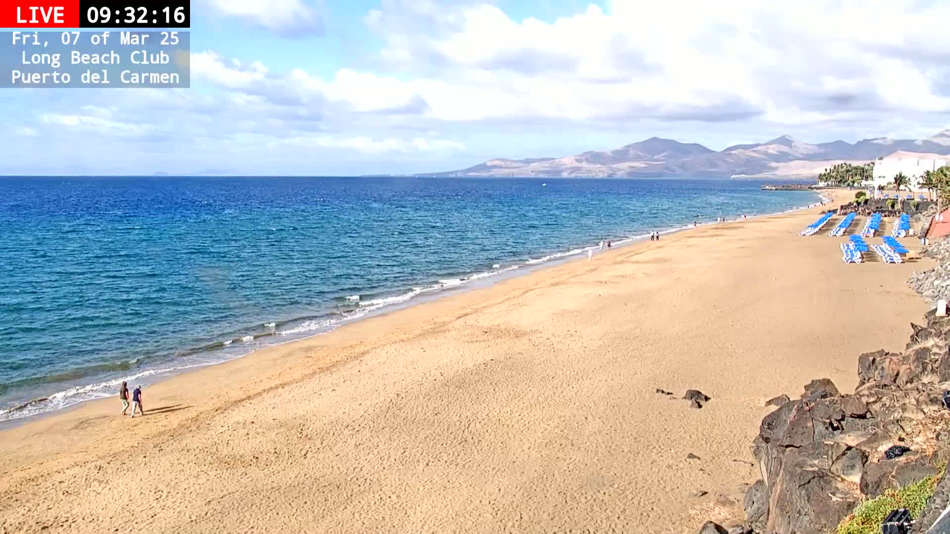 A sandy beach stretches along a calm blue ocean under a partly cloudy sky, with mountains visible in the distance and a few people walking along the shore.