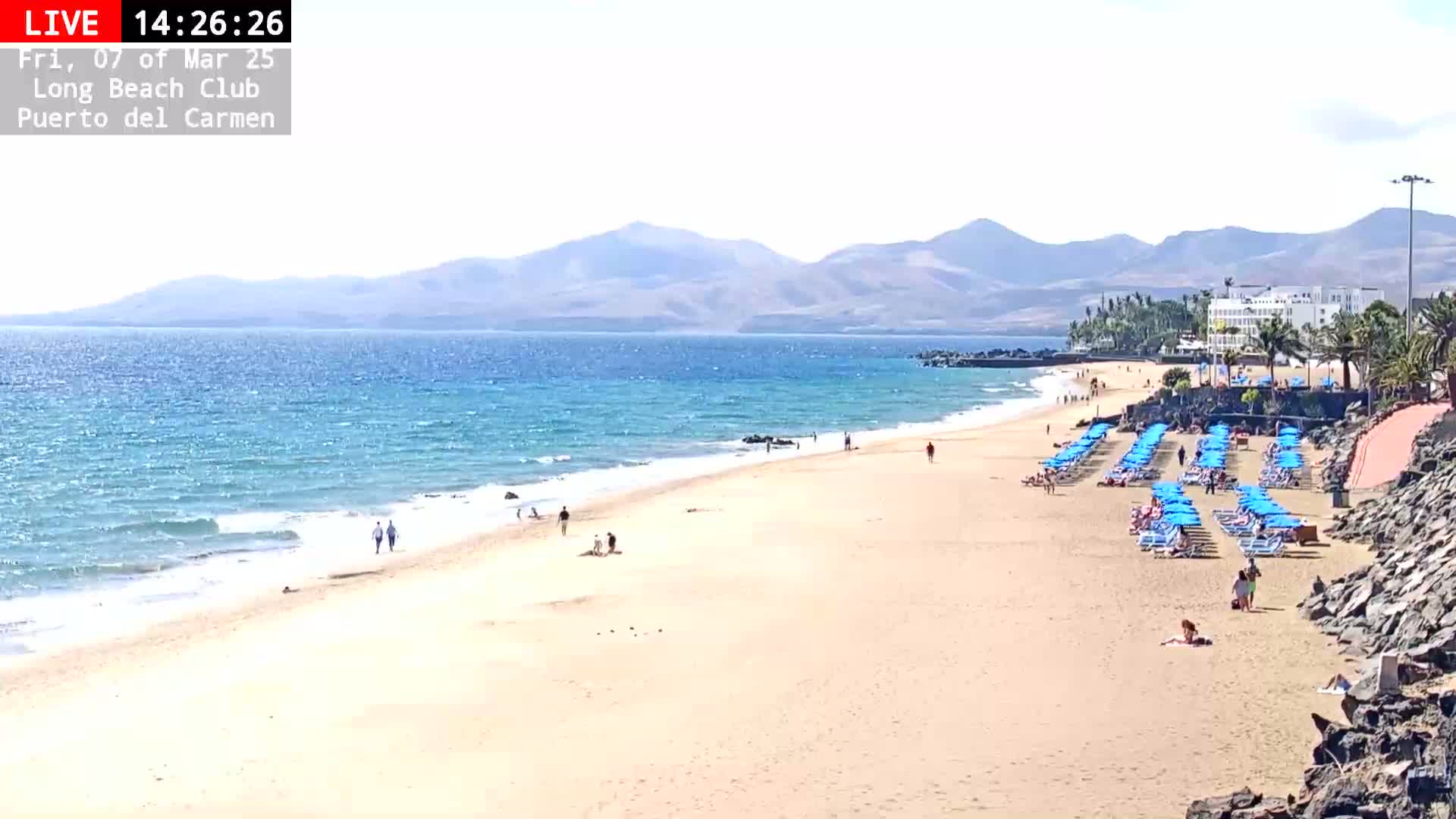A sandy beach with light blue water and rows of blue umbrellas under a sunny sky, backed by distant mountains.