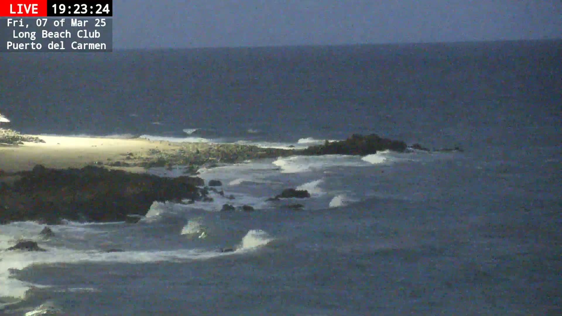 Waves crash against dark rocks near a sandy beach under a twilight sky.