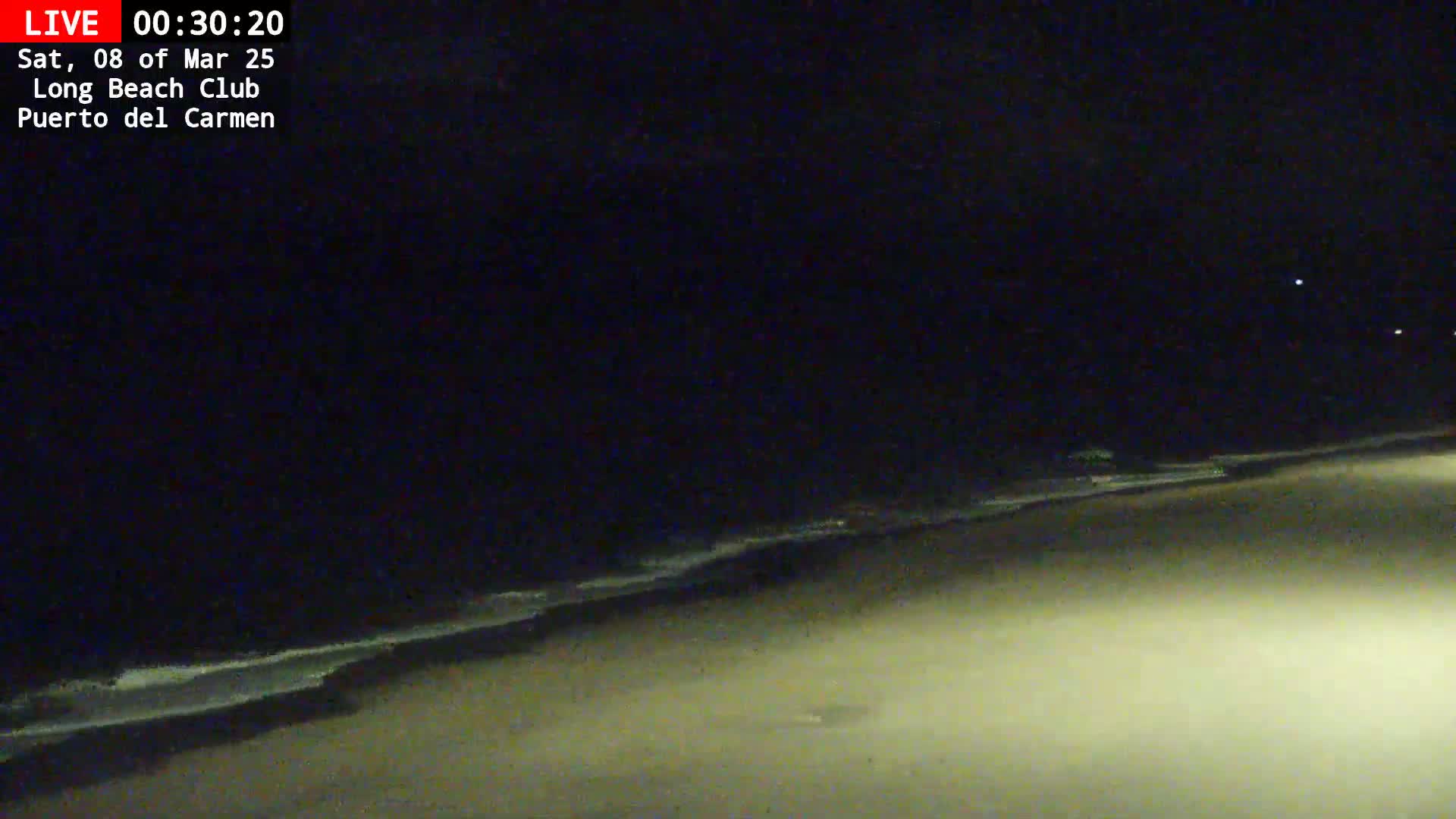 A dark, nighttime beach scene shows gently lapping waves on a sandy shore under a clear sky.