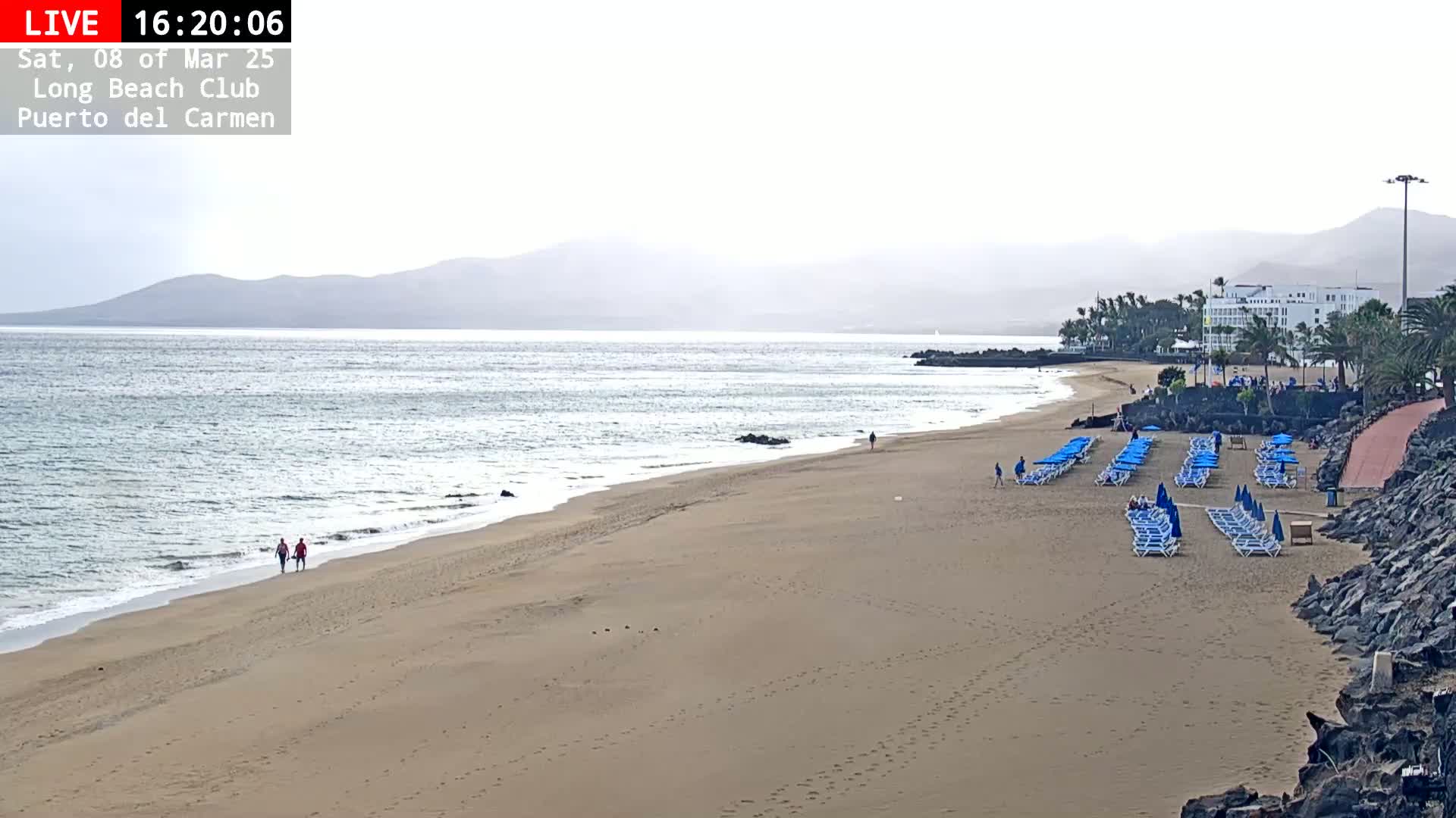 A mostly clear, hazy day shows a sandy beach with rows of blue lounge chairs, a calm ocean, and distant mountains.