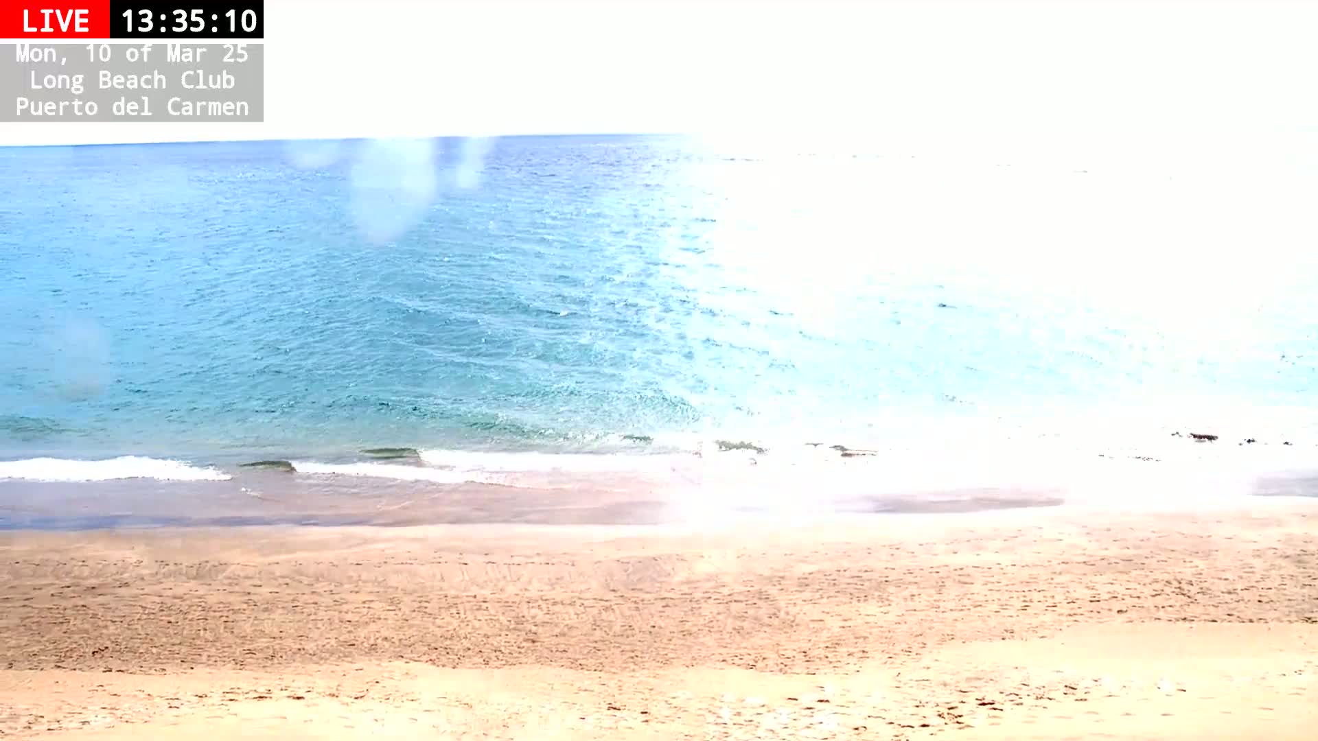A sandy beach slopes down to a calm, light-blue ocean under a bright, mostly clear sky.