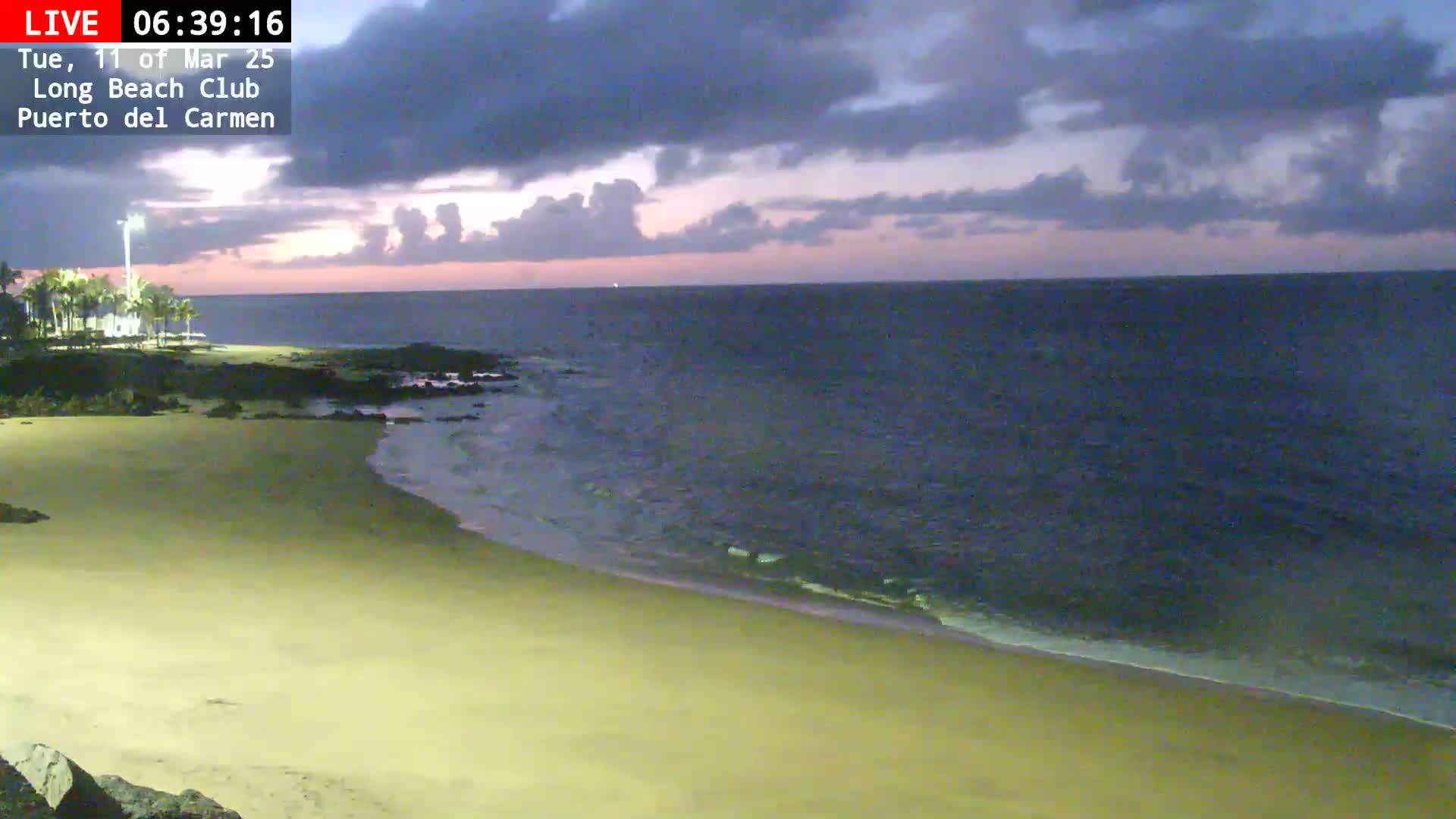 A sandy beach meets a dark-blue ocean under a cloudy, twilight sky.