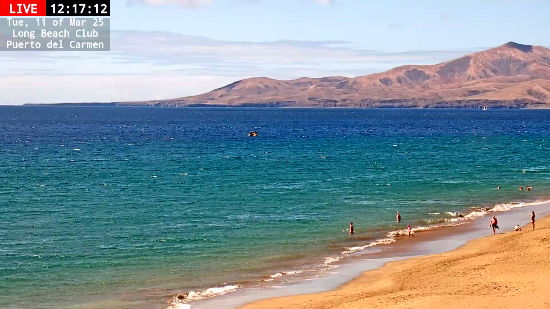 A sandy beach with a few people swimming and sunbathing, with a calm, teal ocean extending to a distant brown mountain range under a mostly clear sky.