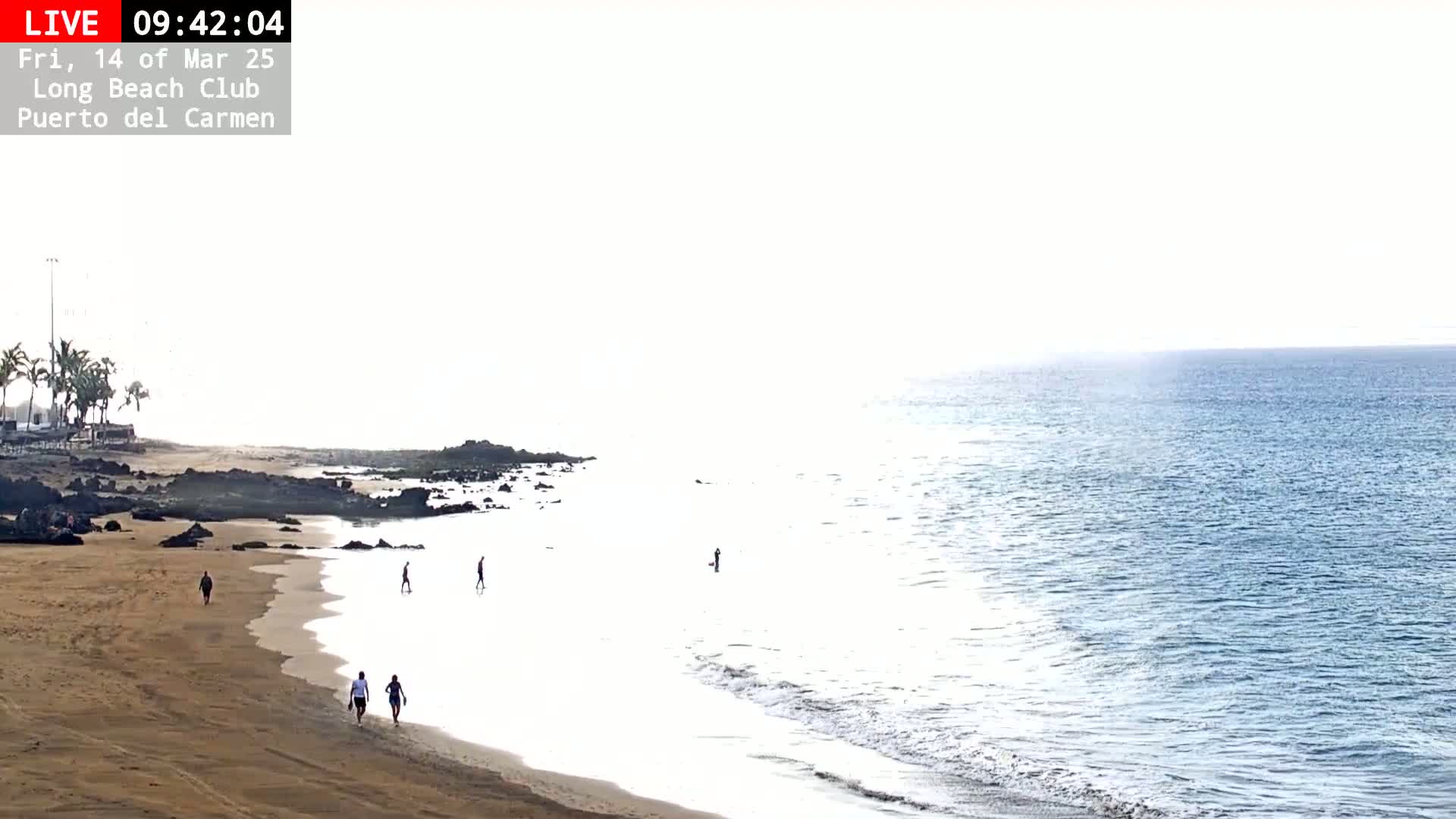 A sandy beach with a few people walking and wading in the calm ocean water under a bright, hazy sky.