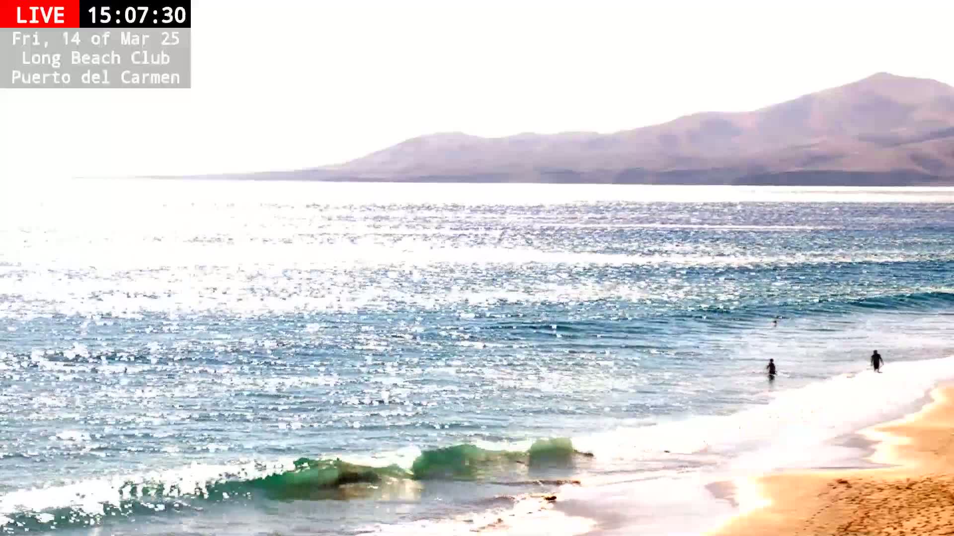 A sandy beach with a few people in the water, gentle waves lapping the shore, under a bright, sunny sky with distant mountains visible across a sparkling ocean.