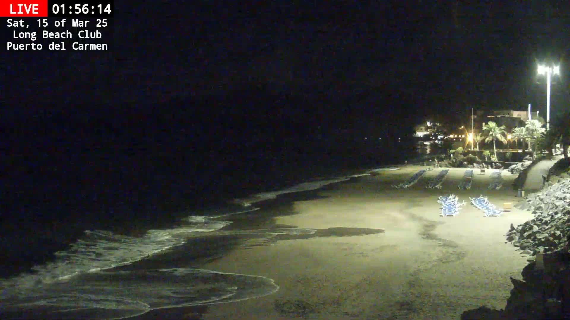 A dark, sandy beach at night shows gentle waves lapping the shore, with rows of empty lounge chairs and a dimly lit building in the background.