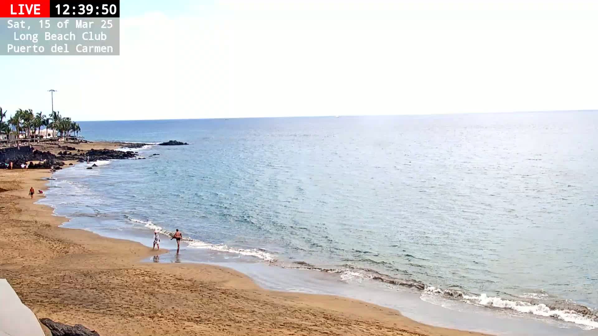 A mostly sunny day at a sandy beach with calm, light blue water and a few people wading near the shore.
