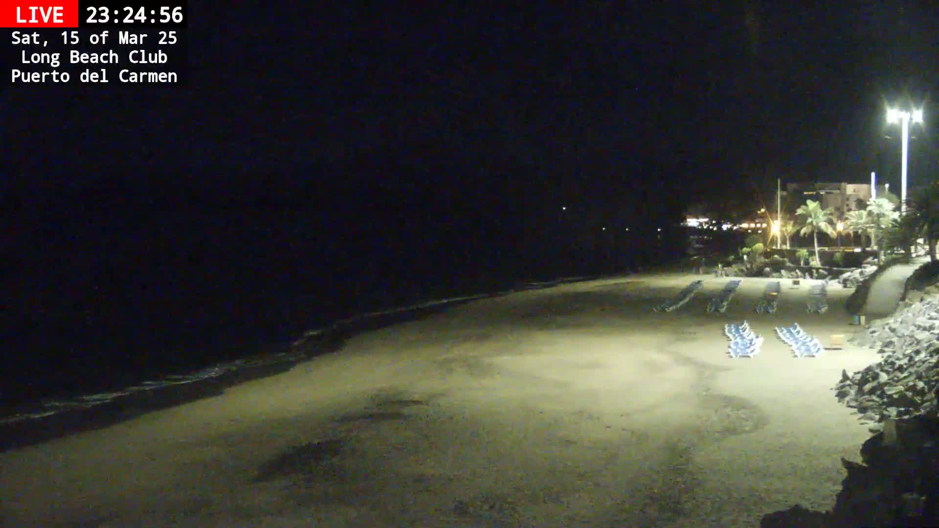 A dark, sandy beach at night shows rows of empty lounge chairs near a rocky shoreline under a clear, dark sky.