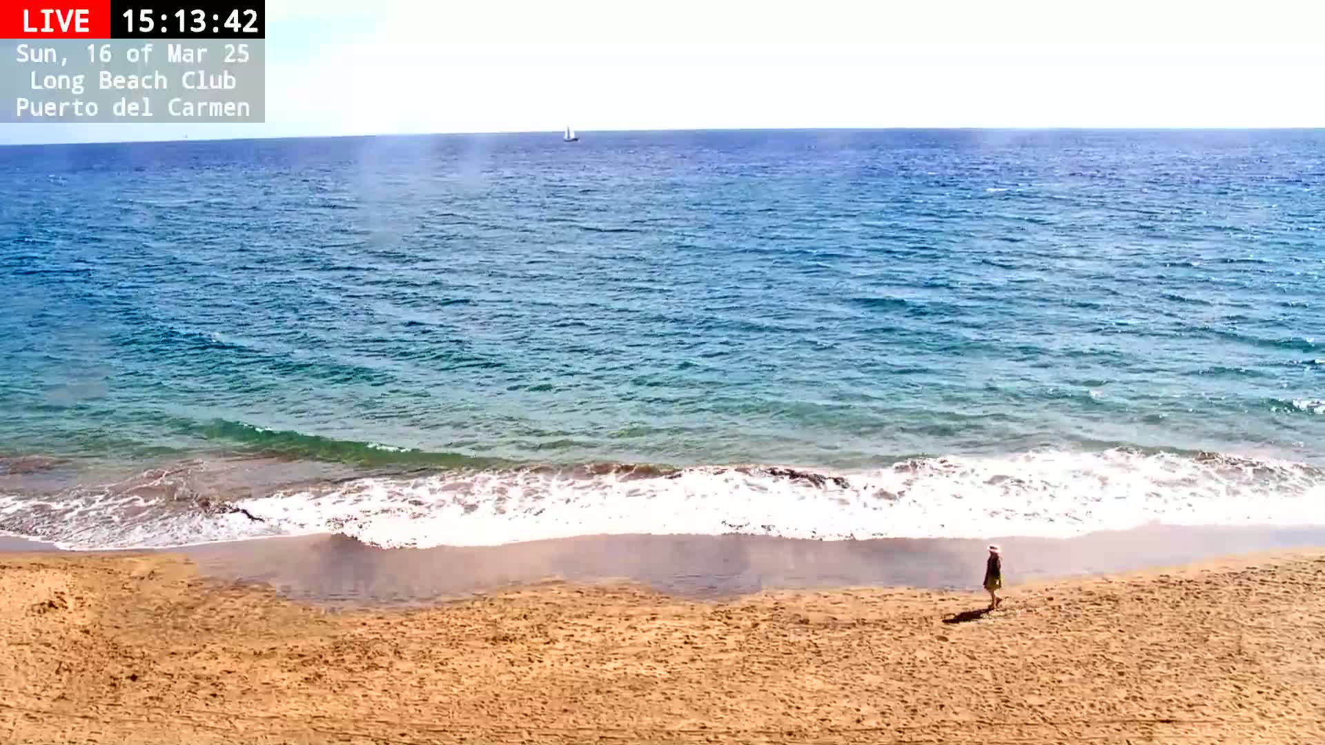 A person walks along a sandy beach with gentle waves lapping the shore on a sunny day.