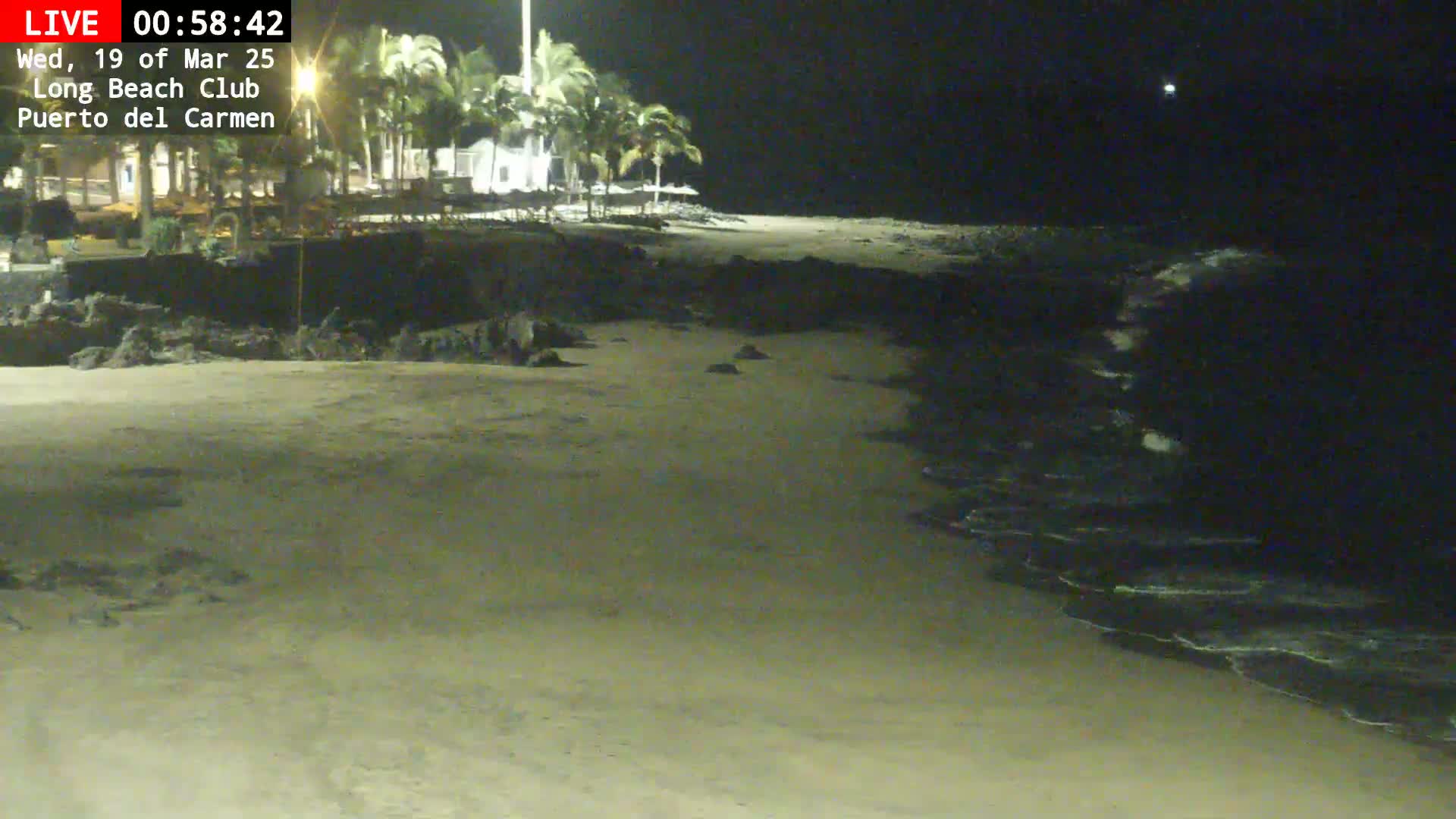 A sandy beach at night with gentle waves lapping the shore, palm trees and buildings visible in the distance under a dark sky.