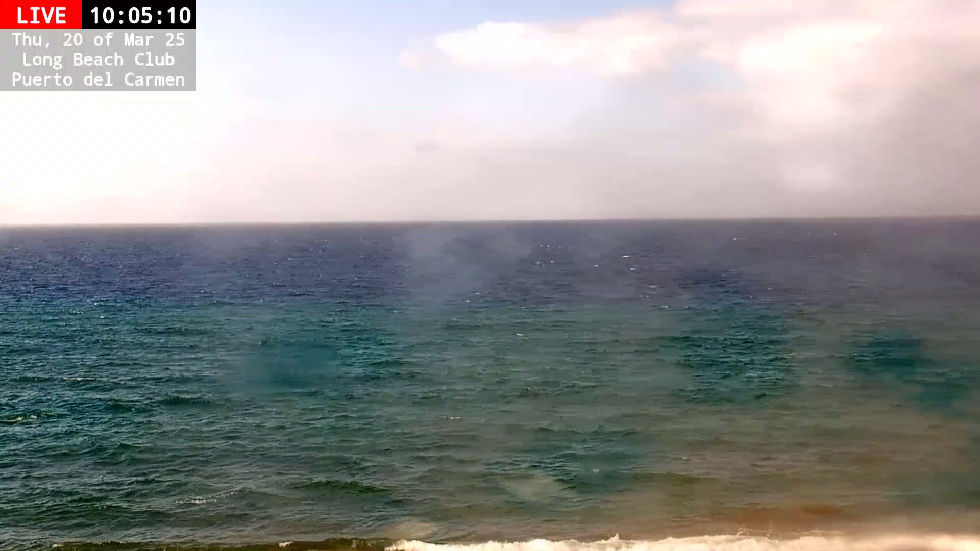 A mostly sunny day shows a wide expanse of dark teal ocean with small waves rolling toward a sandy shore under a partly cloudy sky.