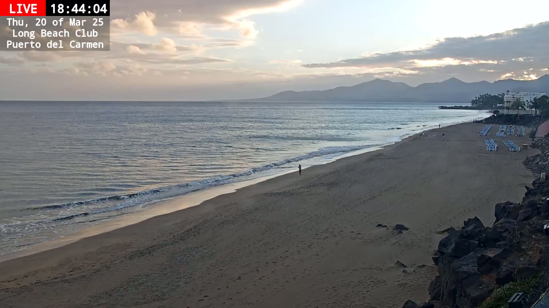 A sandy beach with calm waves under a partly cloudy sky, backed by distant mountains.