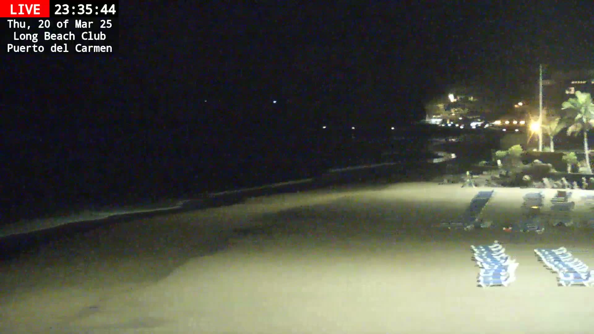 A dark, nighttime beach scene shows a sandy shore with rows of empty lounge chairs, a calm ocean, and distant lights from buildings.