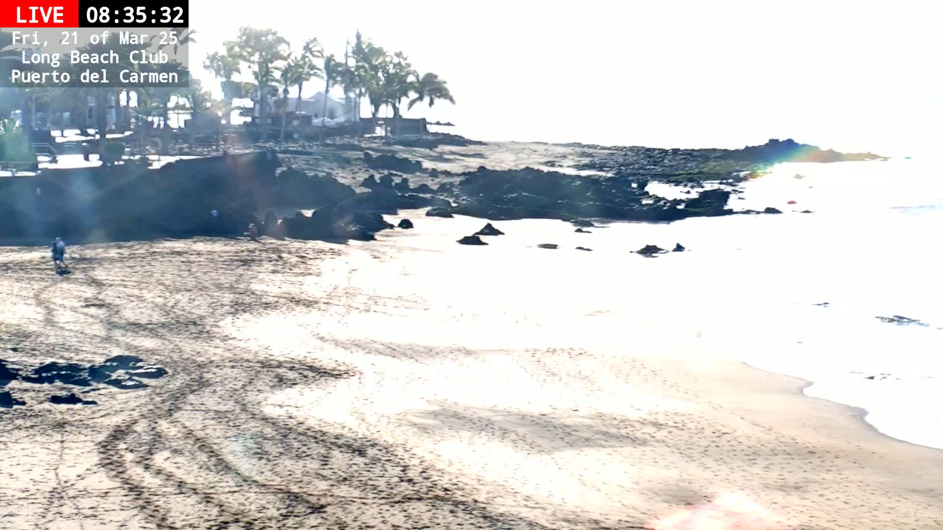 A sandy beach with dark rocks and a few people stretches toward a calm ocean under a bright, hazy sky.