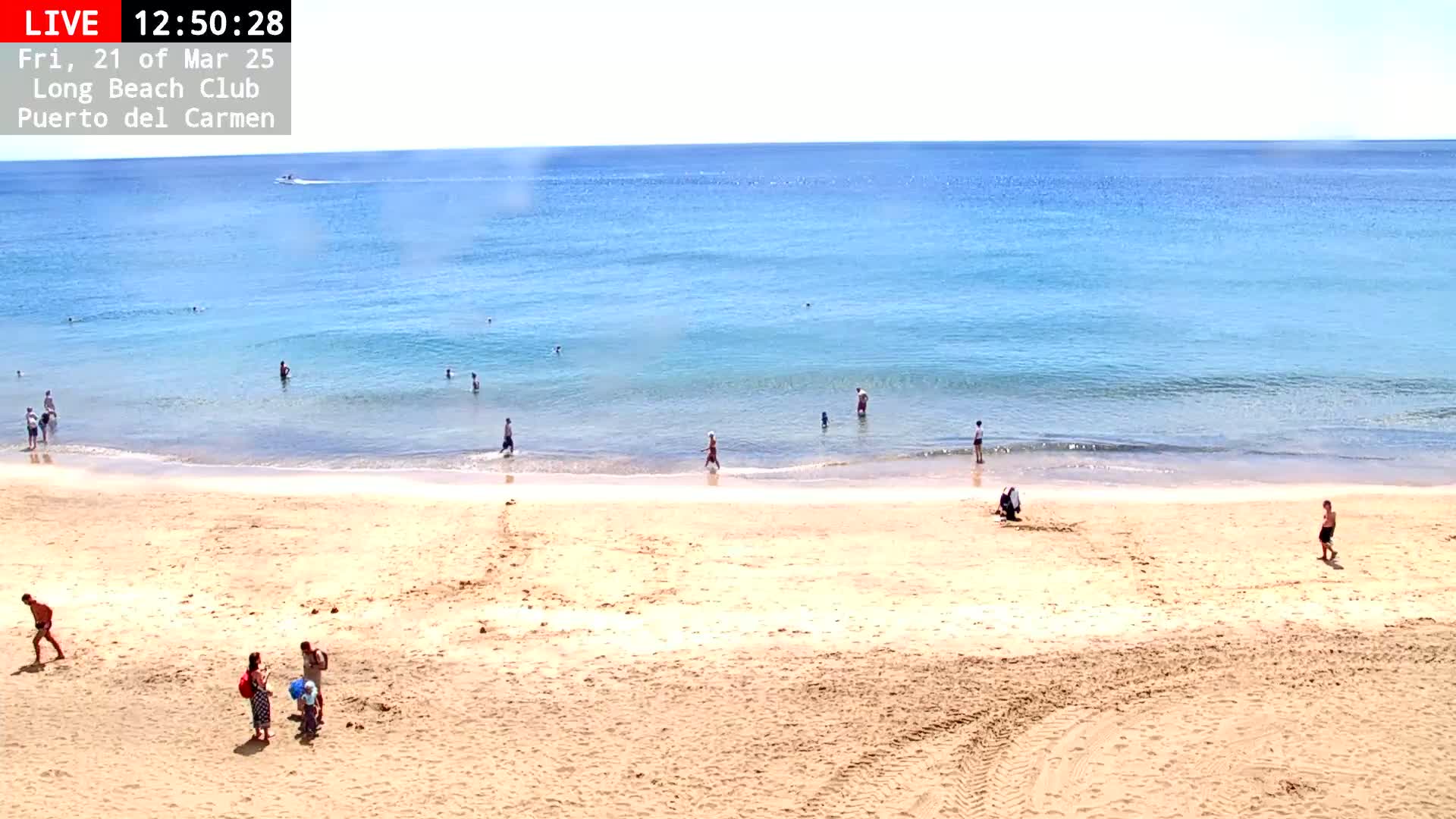 A sandy beach with a few people on it and in the calm, blue ocean under a bright, sunny sky.