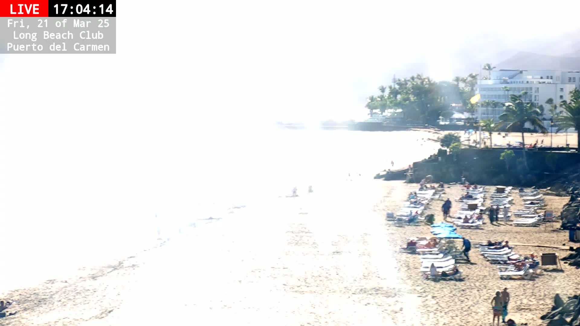 A hazy, sunny beach scene shows a sandy shore with lounge chairs and people, a calm ocean, and a hotel in the background.