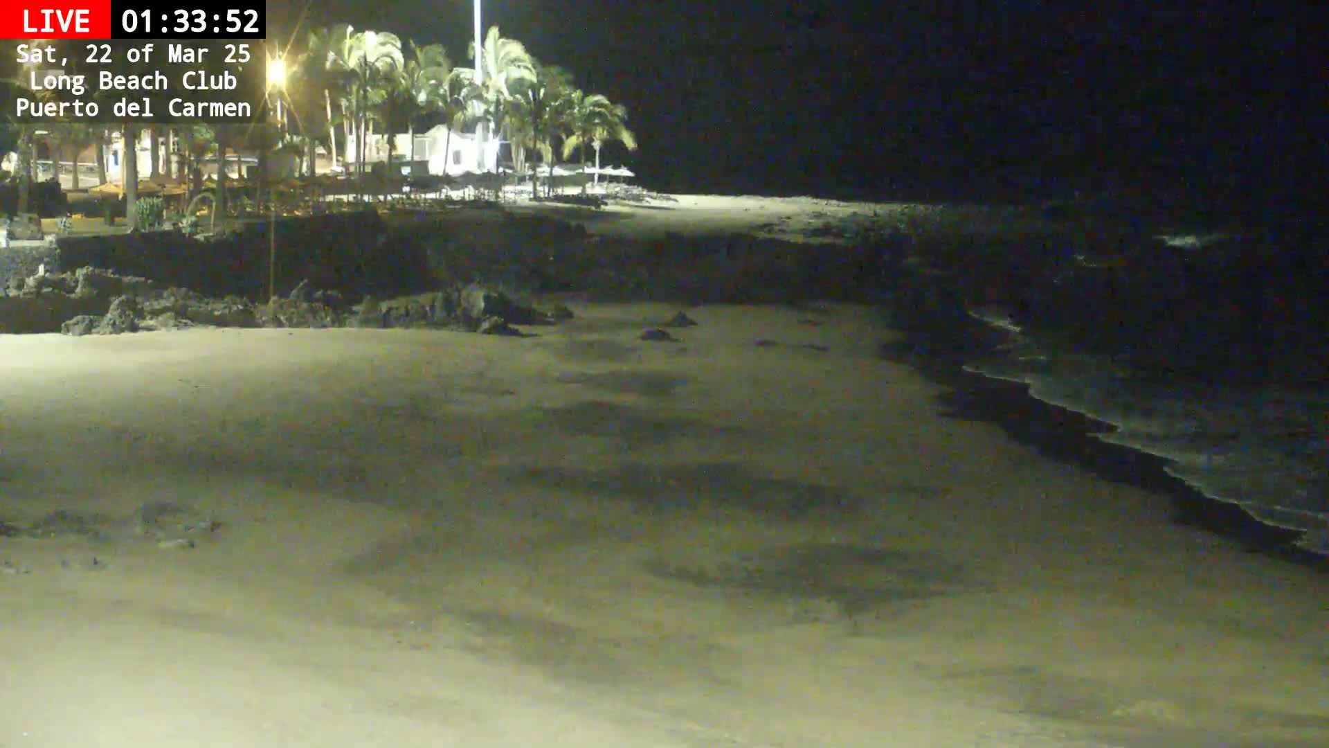 A nighttime beach scene shows a sandy shore with gentle waves lapping at the edge, palm trees and buildings visible in the background under a dark sky.