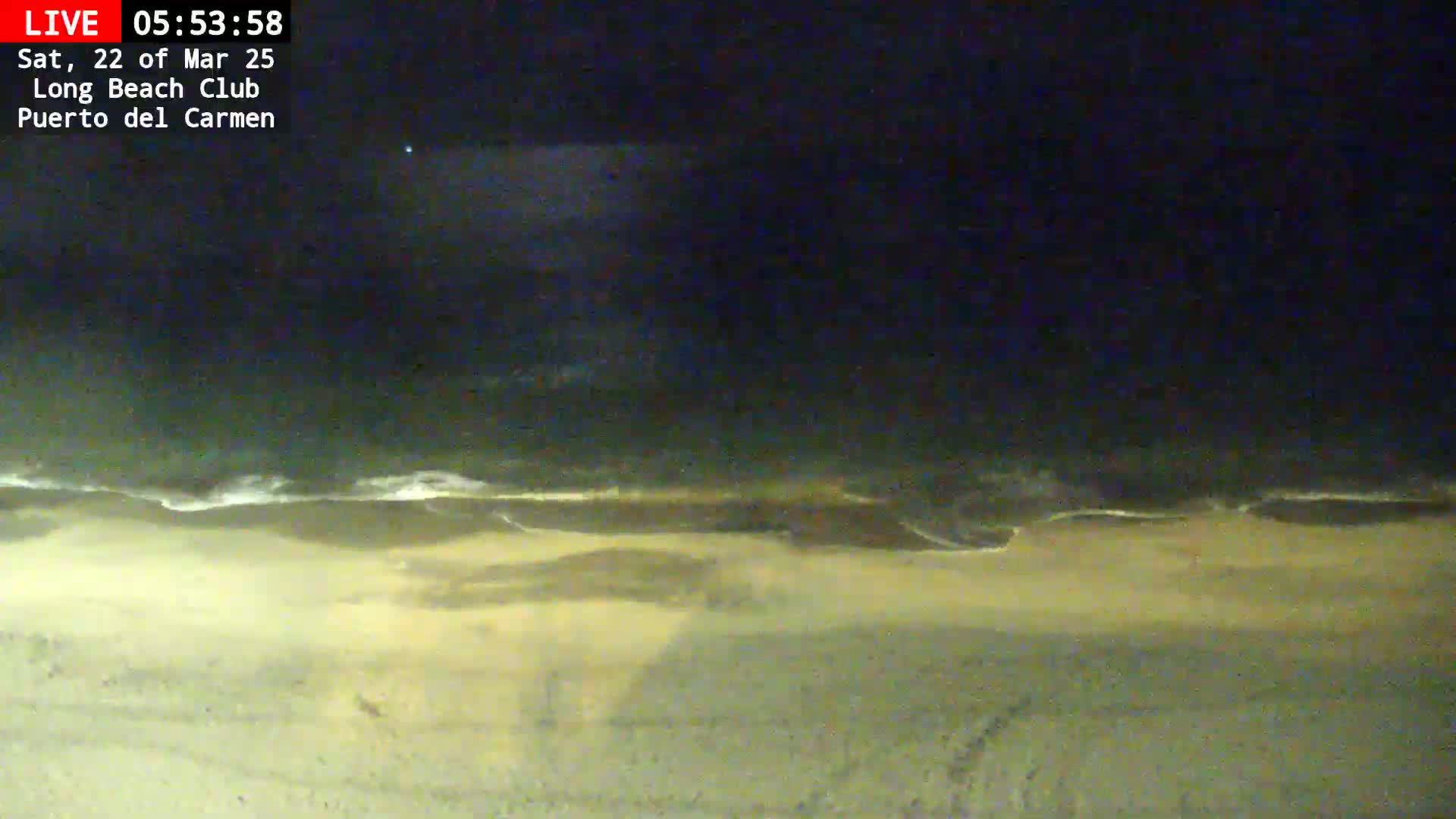 A nighttime, high-angle view shows a sandy beach with gentle waves lapping the shore under a dark sky.