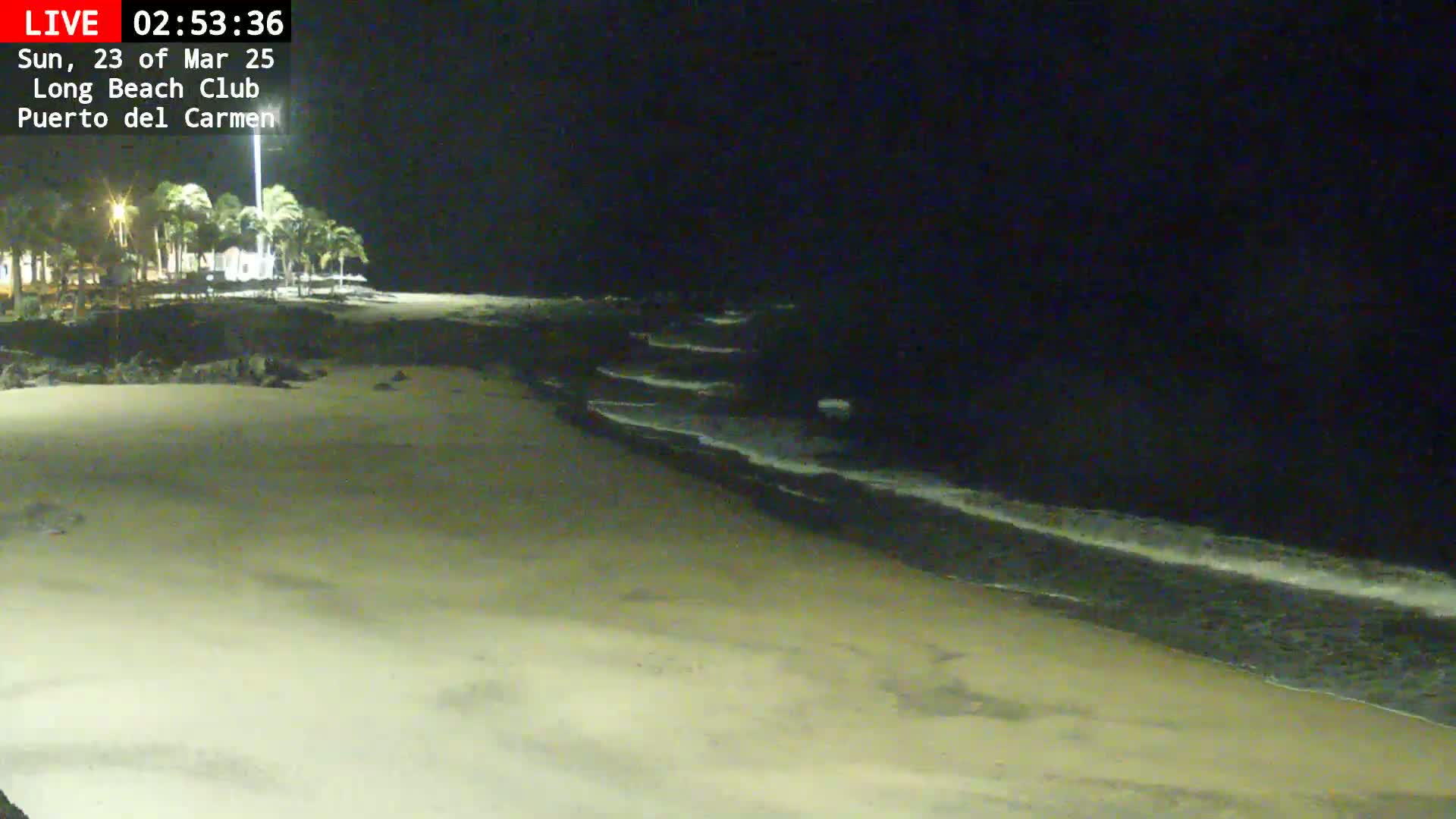 A nighttime view shows a sandy beach with gentle waves lapping the shore under a dark sky.