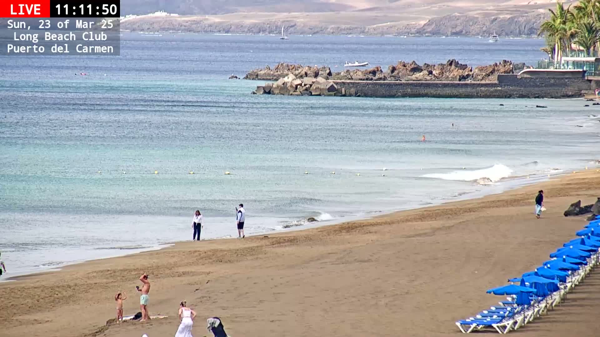 A sandy beach with calm, light-blue water and a few people is seen on a sunny day, with rows of blue beach umbrellas in the foreground and a rocky structure in the midground.