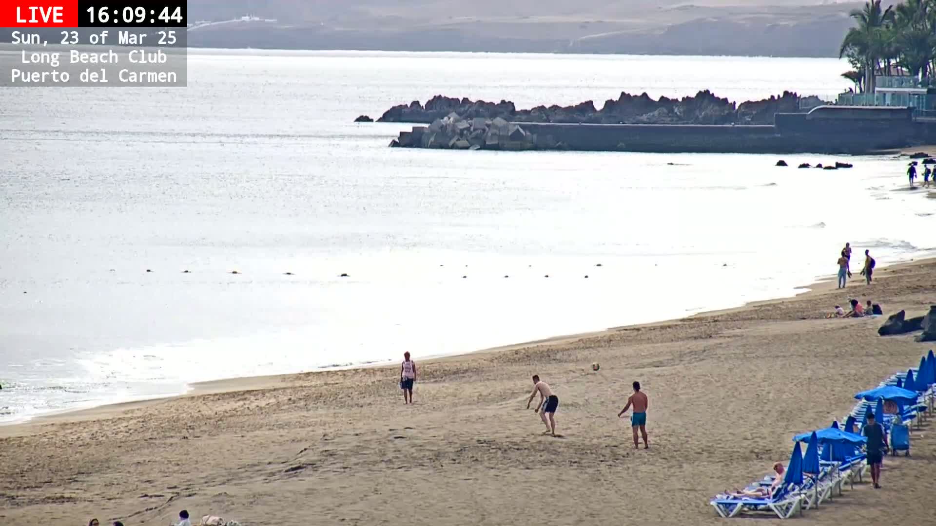 A sandy beach with calm ocean water and a few people playing a game on the sand, under a mostly clear sky.