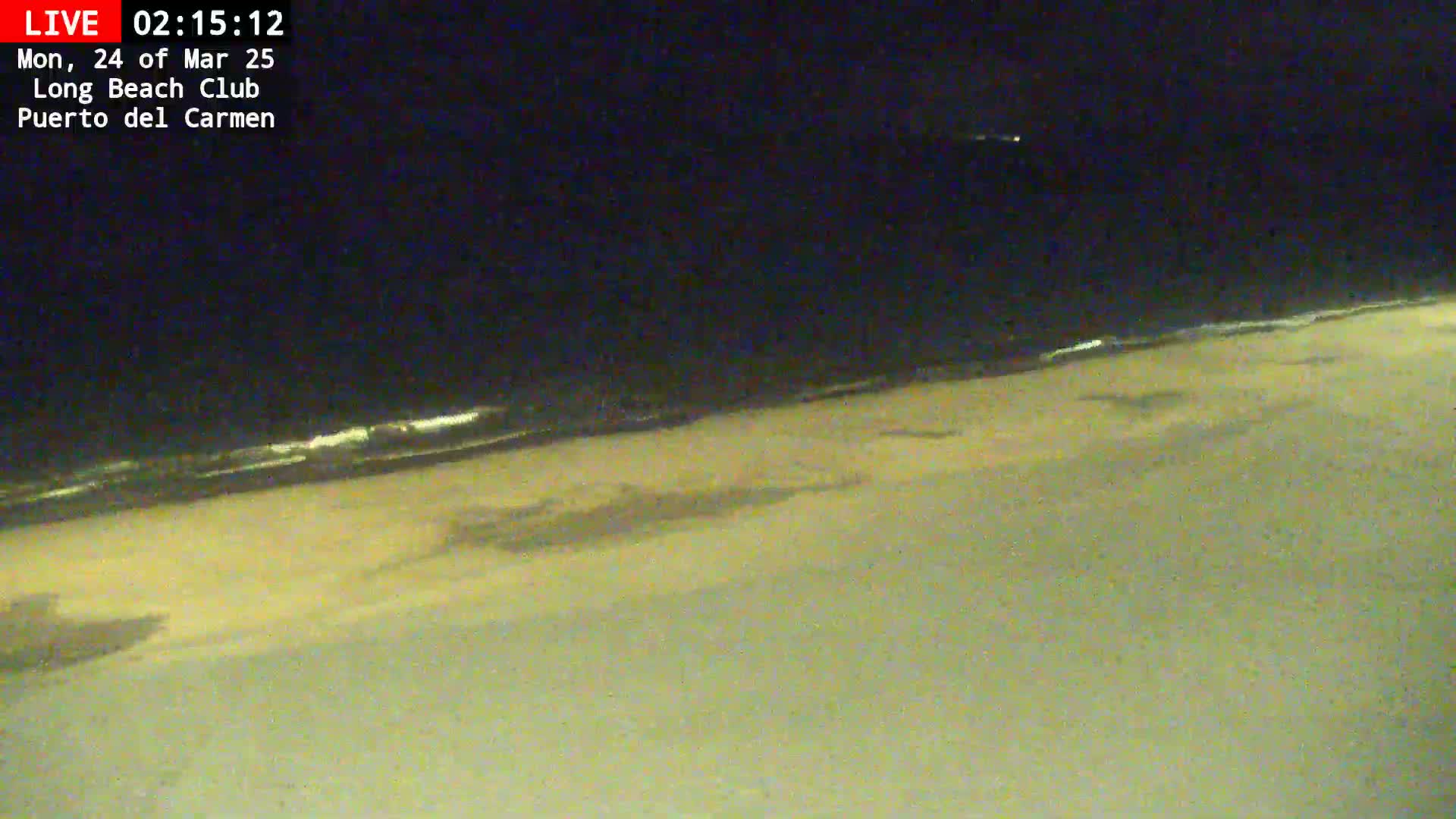 A dark, nighttime beach scene shows a sandy shore with gentle waves lapping at the waterline under a clear, dark sky.